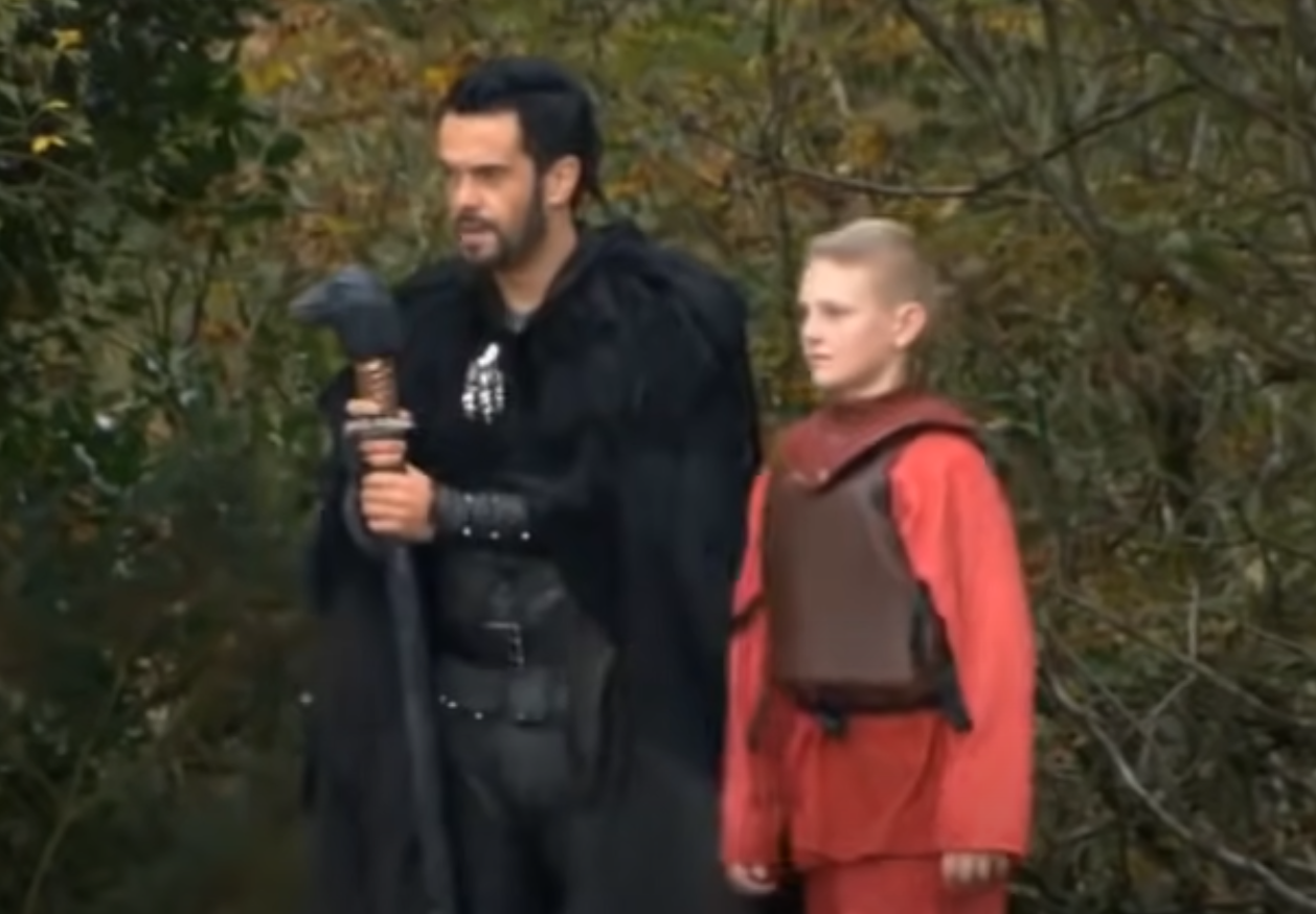 A man in a black cloak holding a raven headed staff and a child in red jumpsuit.