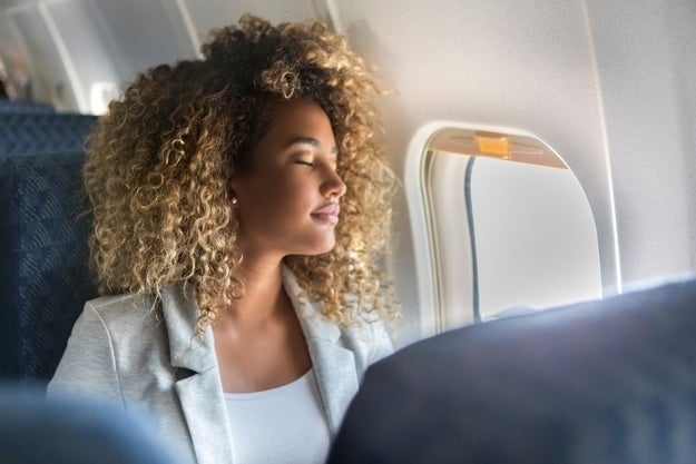 A young woman sits in the window seat of a commercial airliner and leans back with her eyes closed