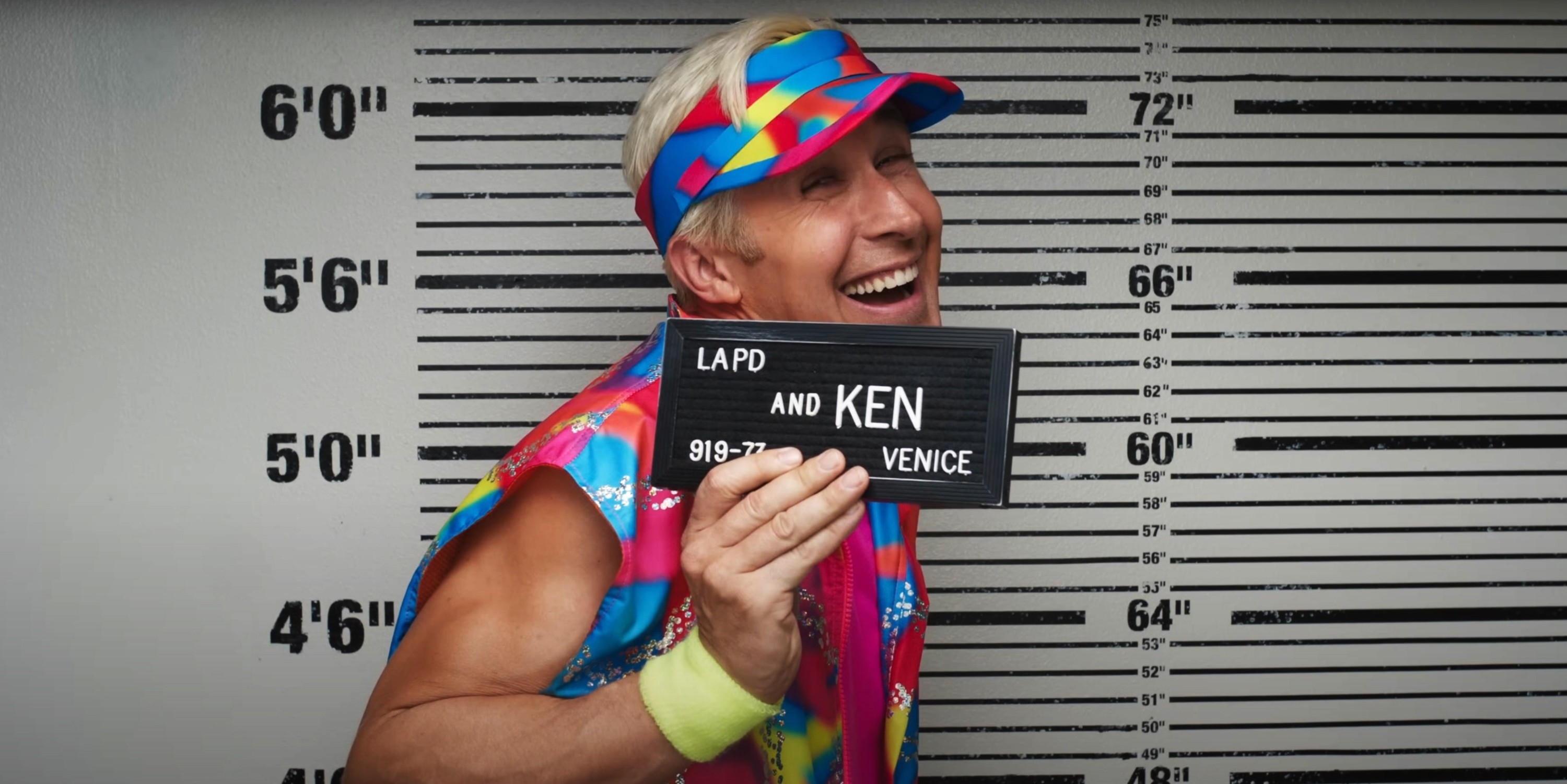 Close-up of Ryan as Ken holding up an LAPD sign and smiling for his mugshot