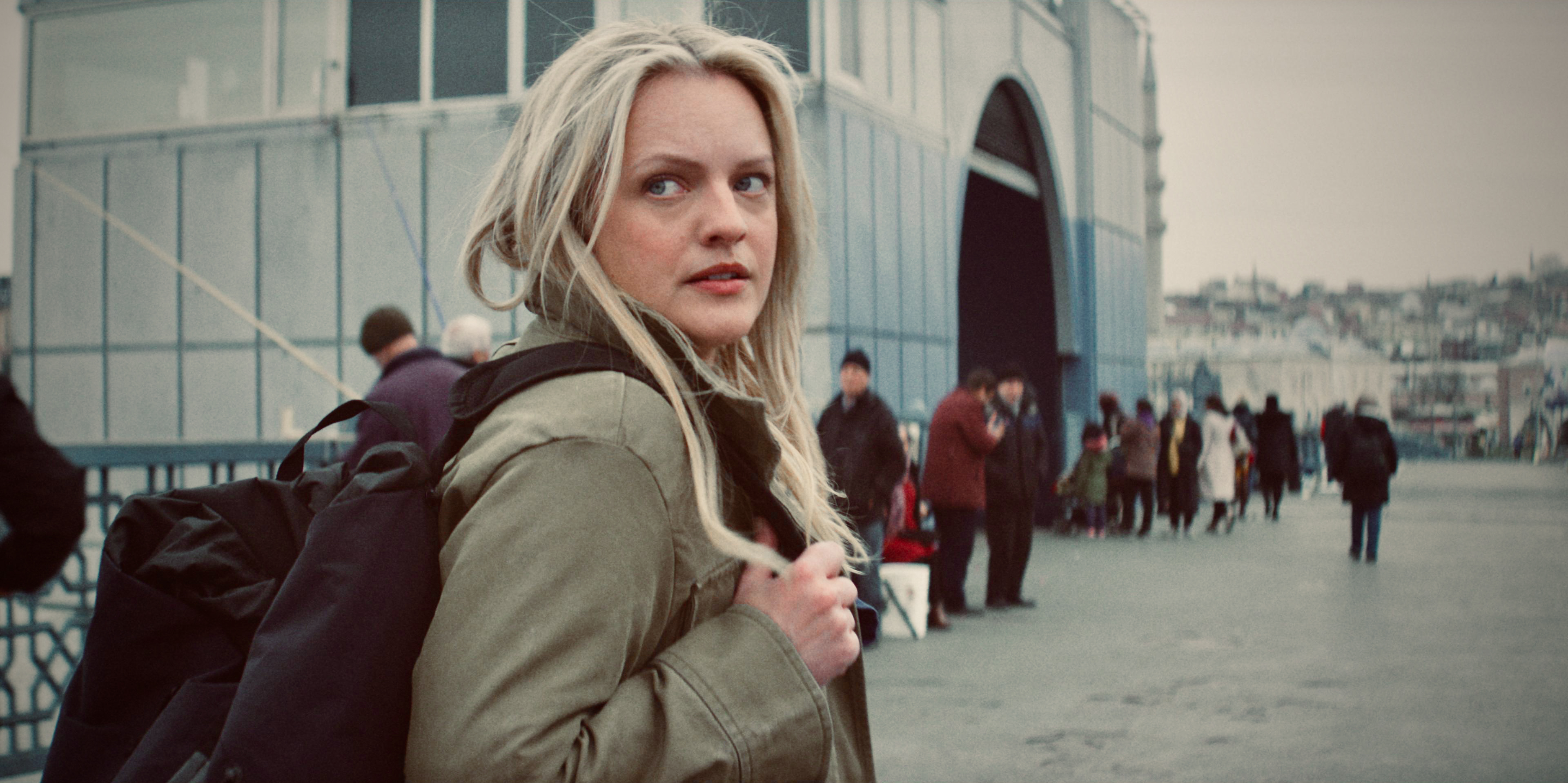 Elisabeth Moss on a crowded street, looking back over her shoulder, a tense expression on her face