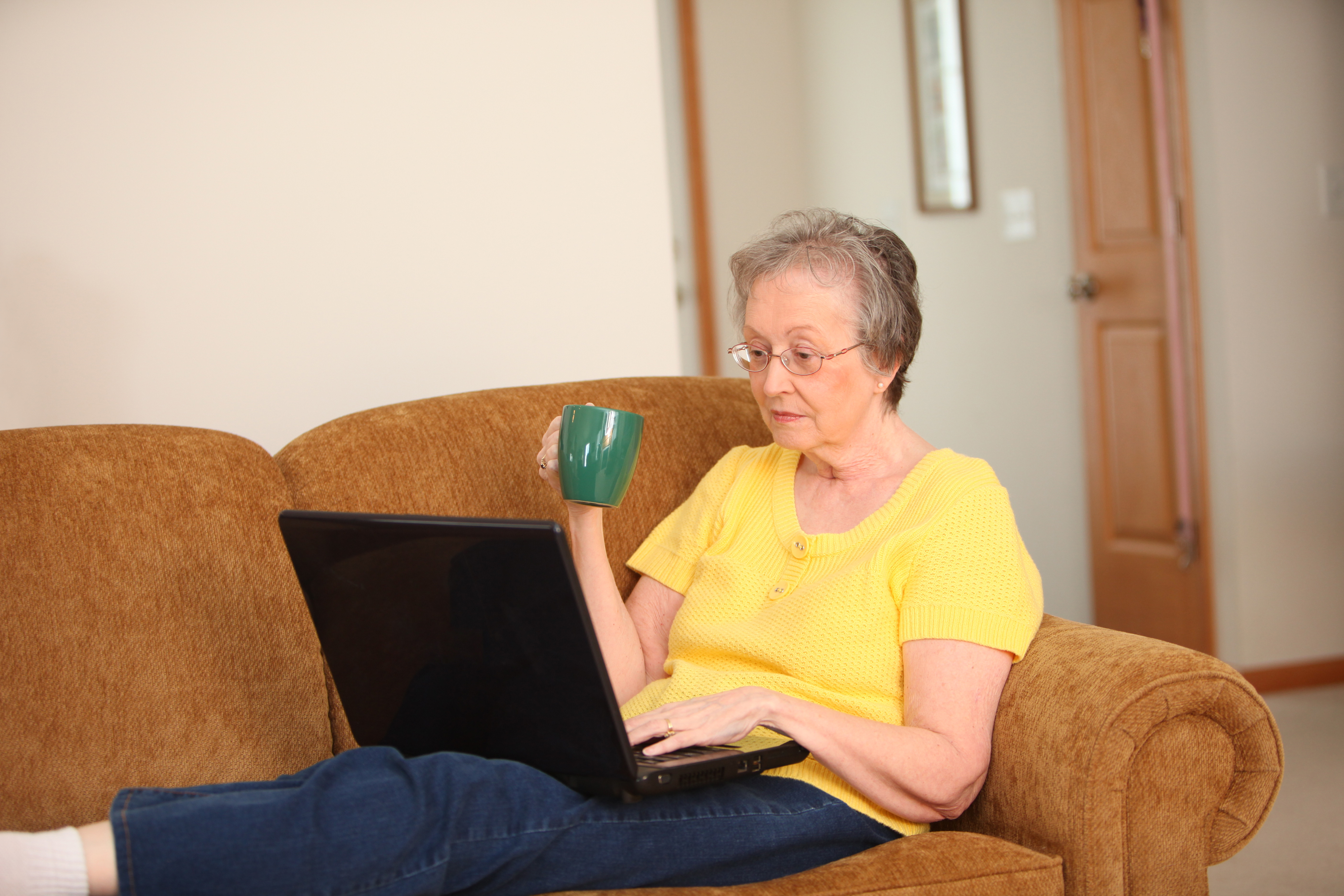 older woman drinking coffee and sitting on the couch with her laptop