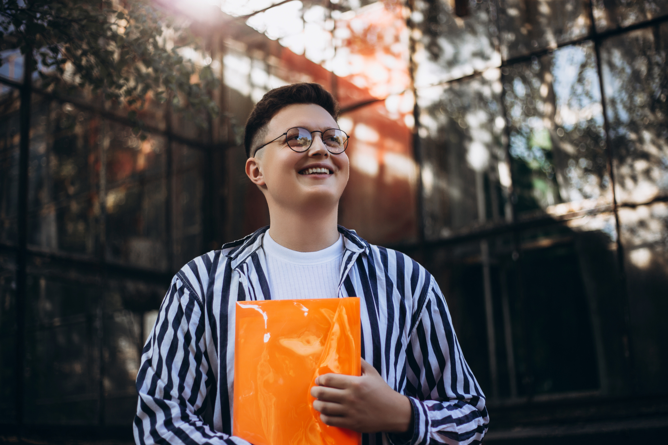 Person holding an orange folder and smiling outdoors, wearing glasses and striped shirt