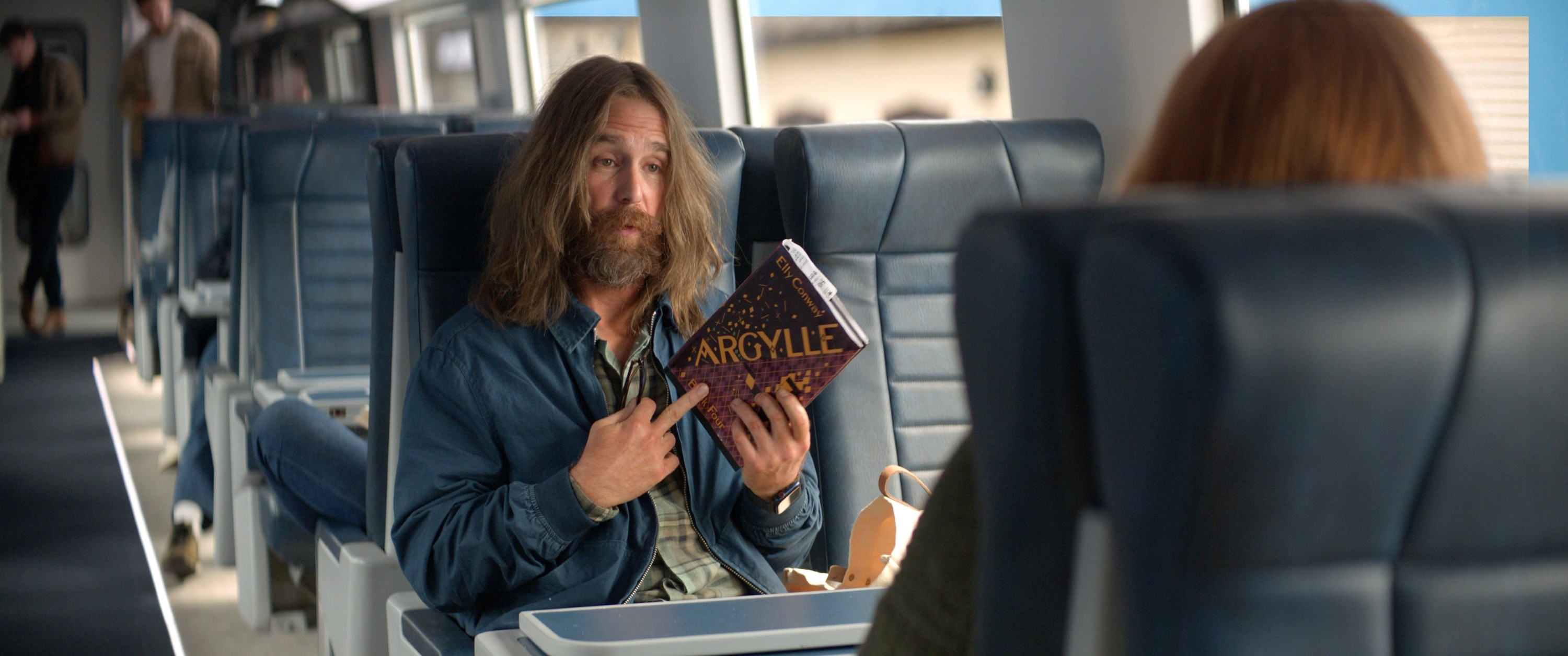 A man with long hair, a beard, and a mustache sitting on a train and holding a copy of Argylle