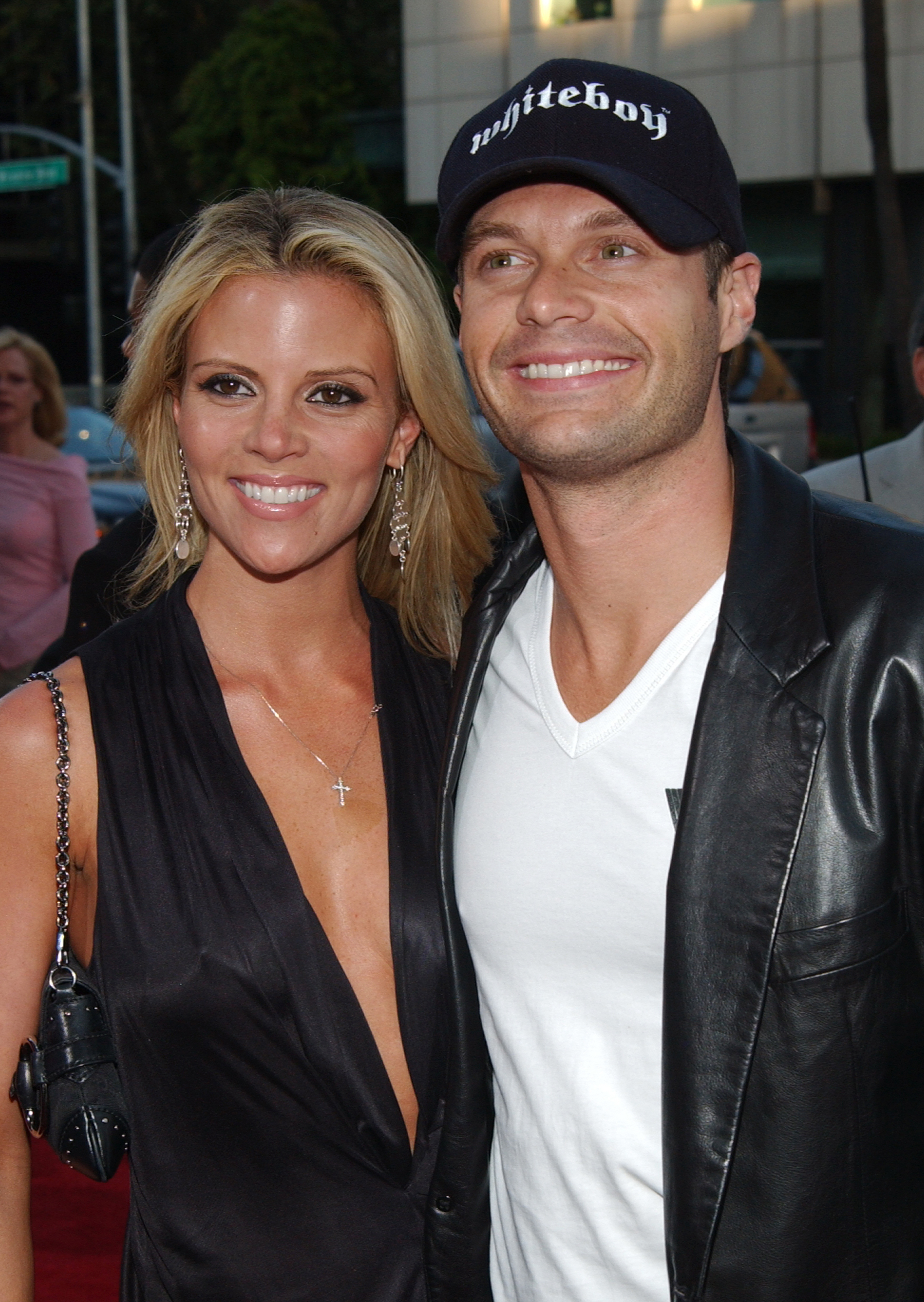 They're smiling on the red carpet; she's wearing a deep-V sleeveless outfit and he's wearing a cap, leather jacket, and shirt