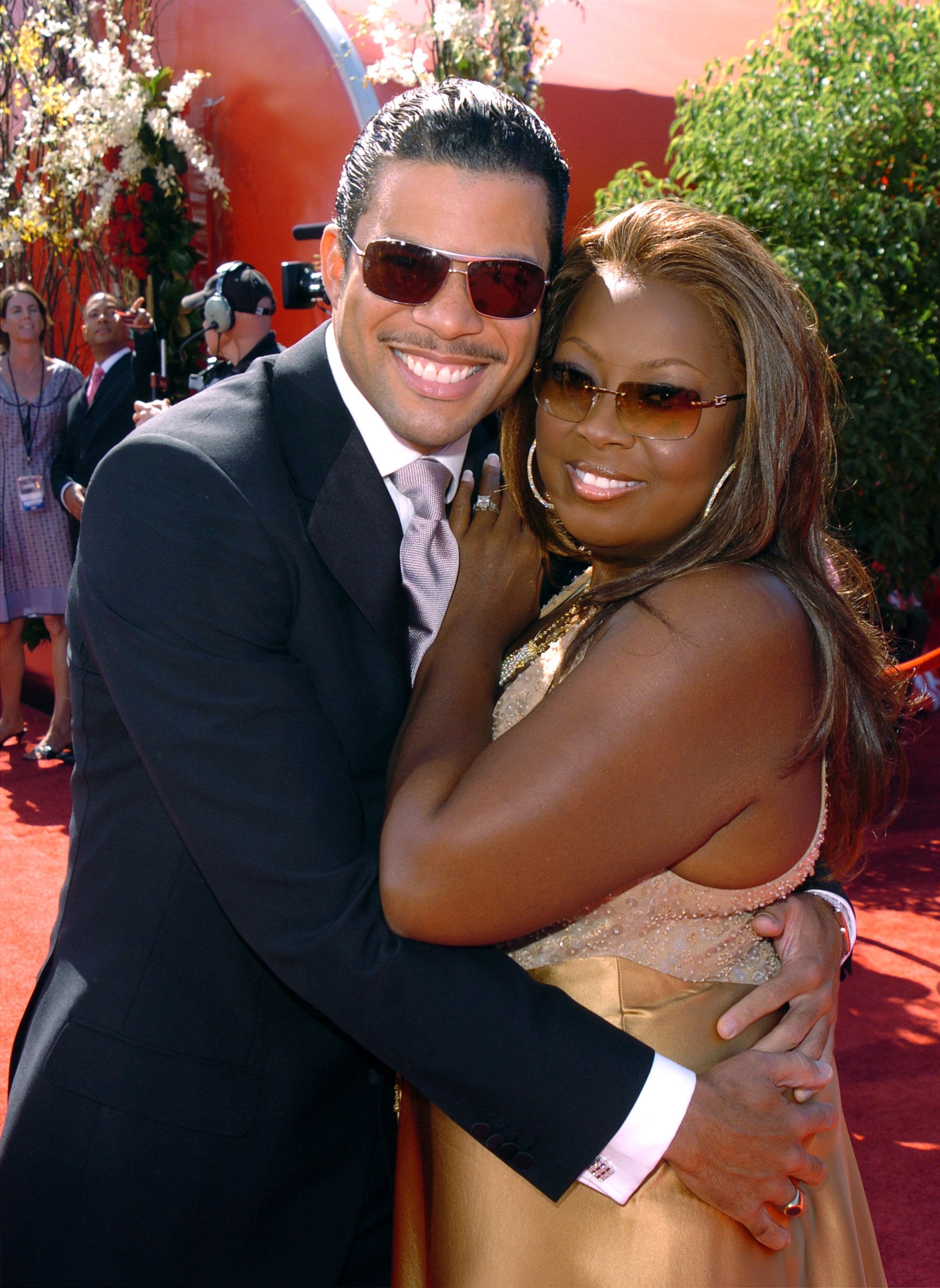 On the red carpet, he's smiling, wearing a suit and tie and sunglasses, and has his arms around her; she's smiling and wearing a sleeveless dress and sunglasses