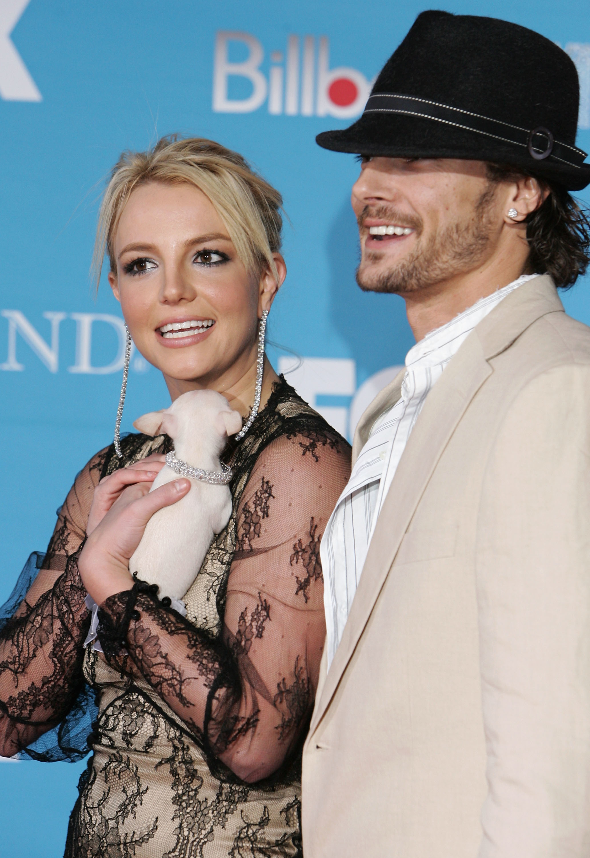 They're smiling at a media event; she's holding a small dog and wearing a lacy, long-sleeved outfit; he's wearing a shirt and suit jacket