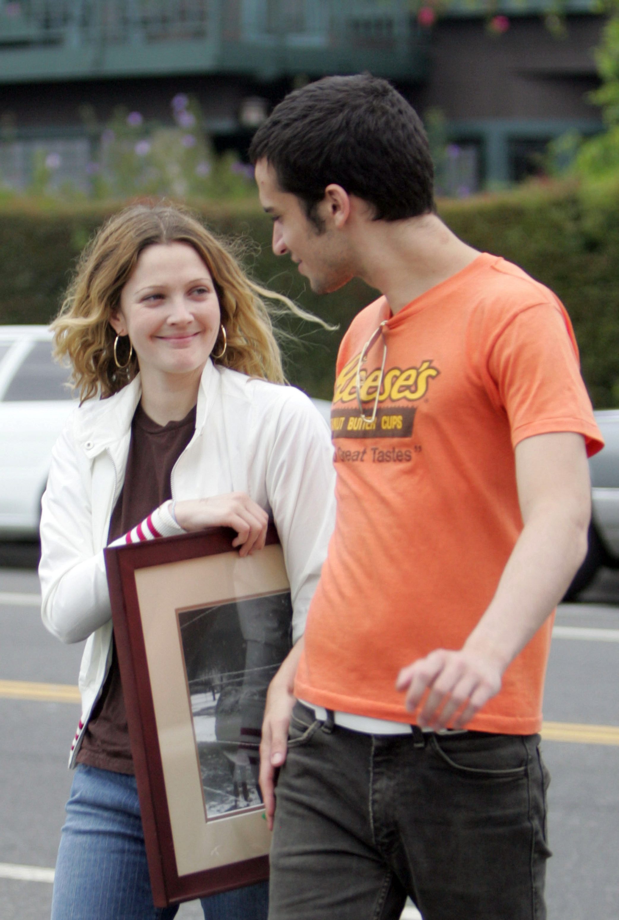 They&#x27;re crossing the street and smiling at each other; she&#x27;s holding a framed photo and wearing a sweater and jeans; he&#x27;s in jeans and a Reese&#x27;s T-shirt