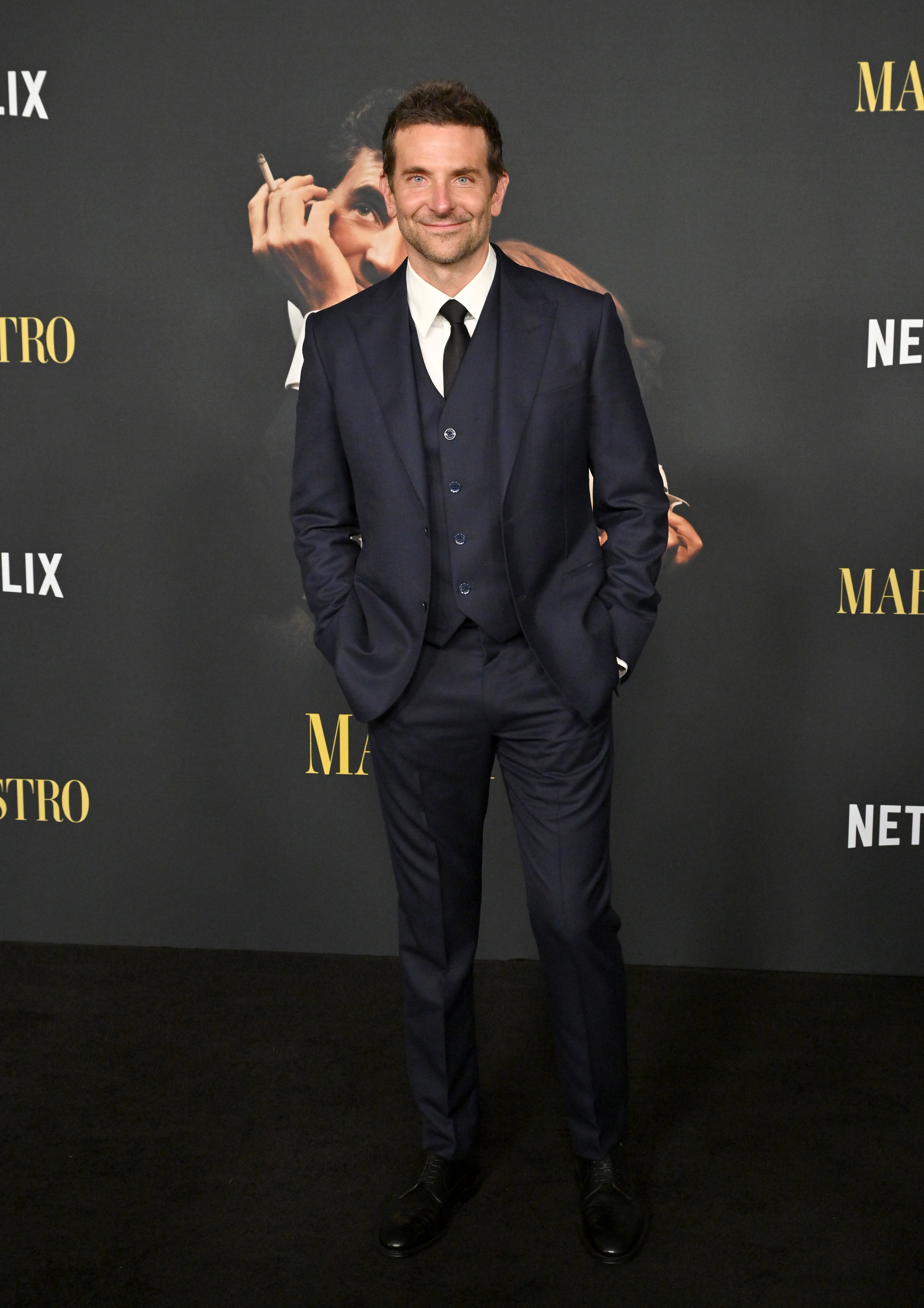 Bradley Cooper stands smiling in a tailored navy suit with a tie, at a Netflix event for &quot;Maestro&quot;