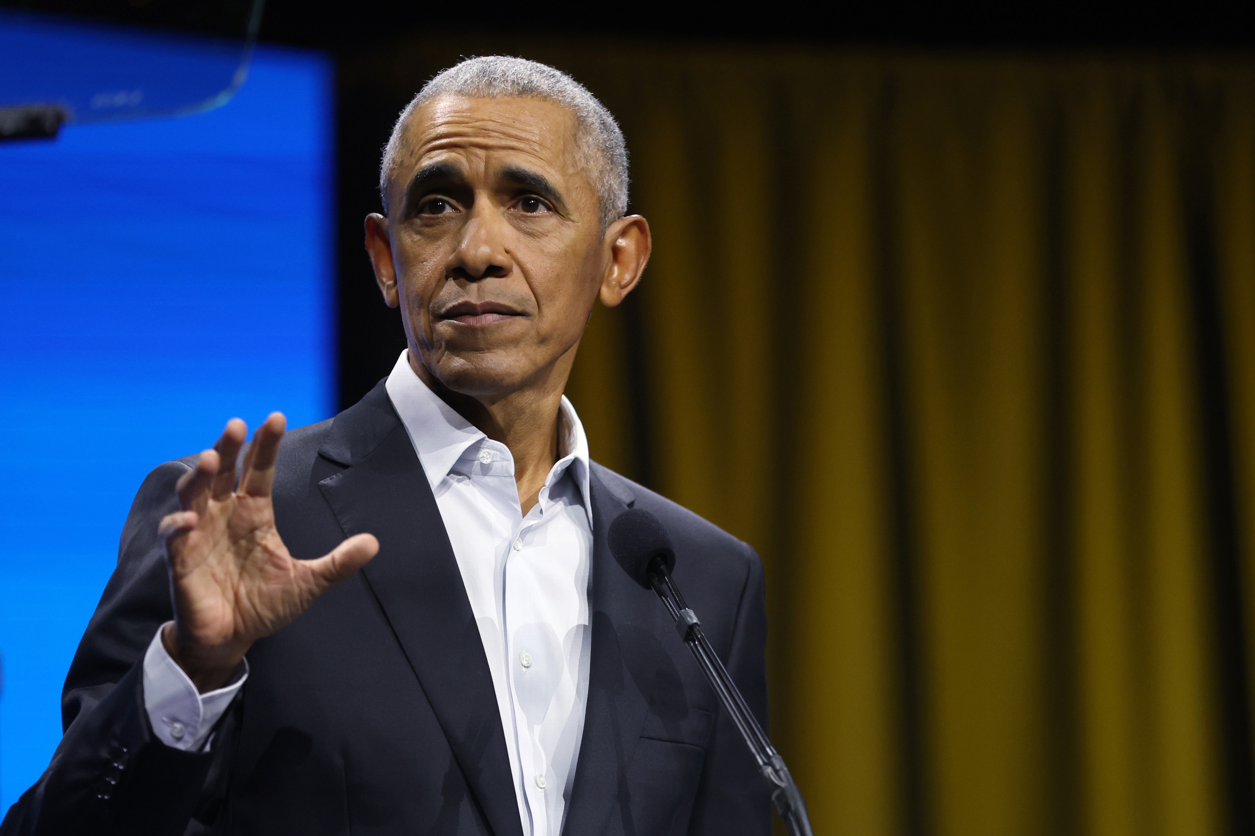 Barack Obama speaking at an event, wearing a suit without a tie, gesturing with his right hand