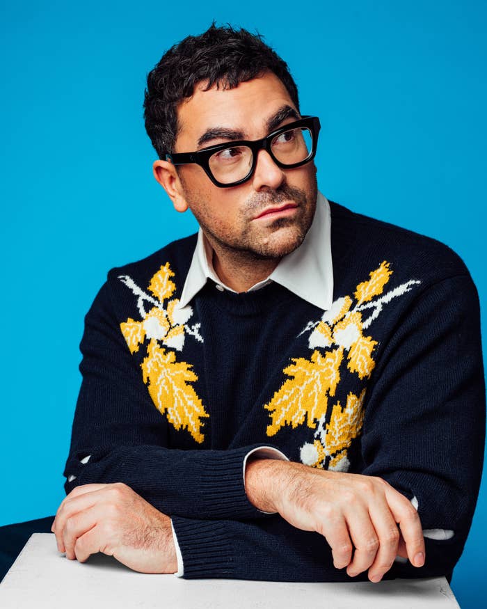 A closeup of Dan Levy in a sweater with his arms crossed on a counter