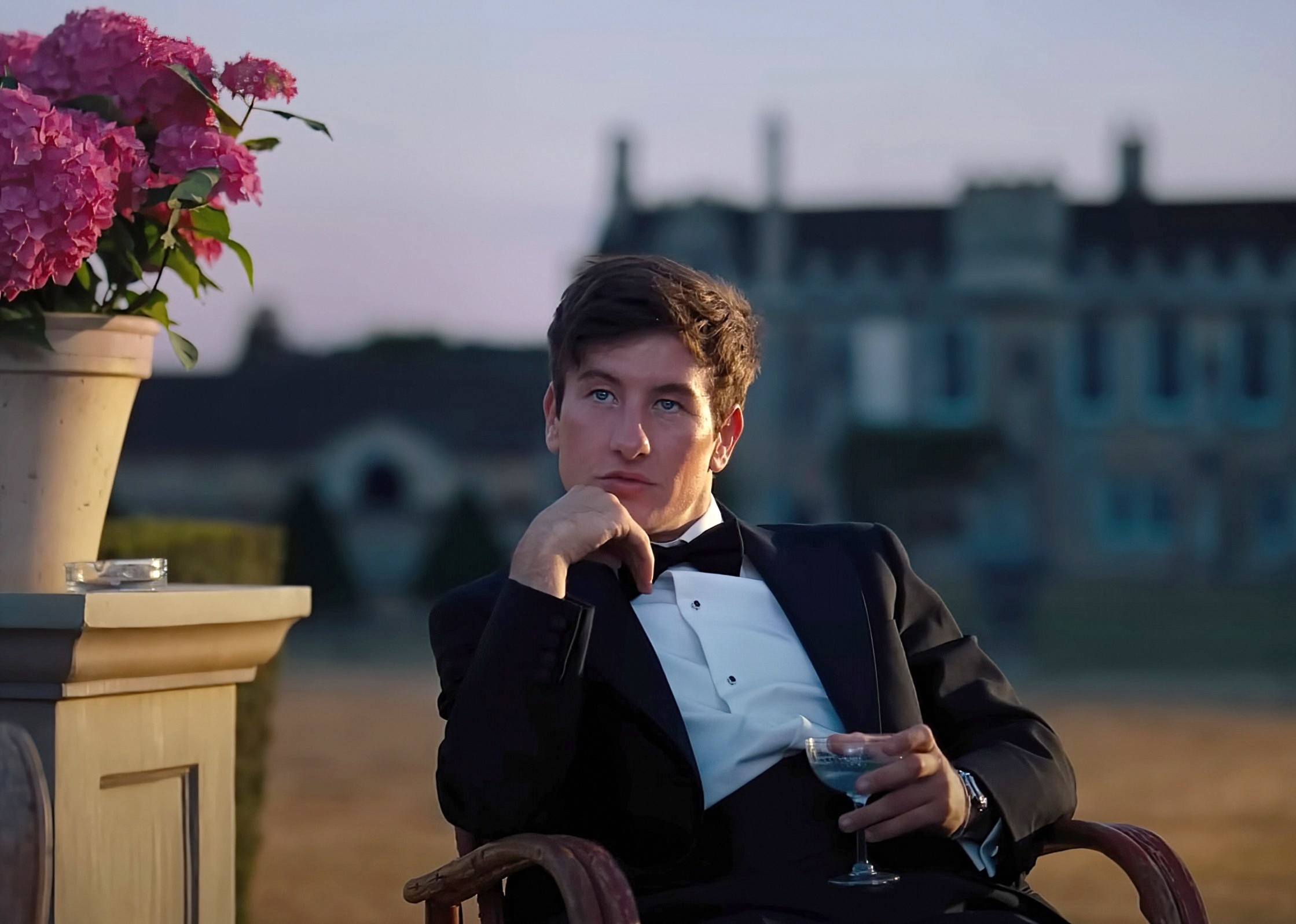 Oliver sits on a chair outside while wearing a tuxedo in a scene from &quot;Saltburn&quot;