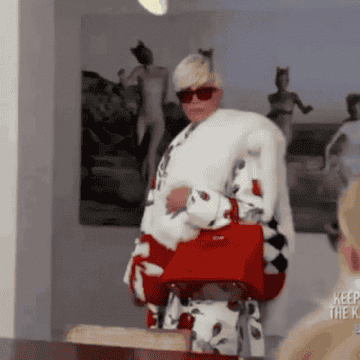 Kris Jenner walking, wearing blonde wig, white fur shrug, and red purse