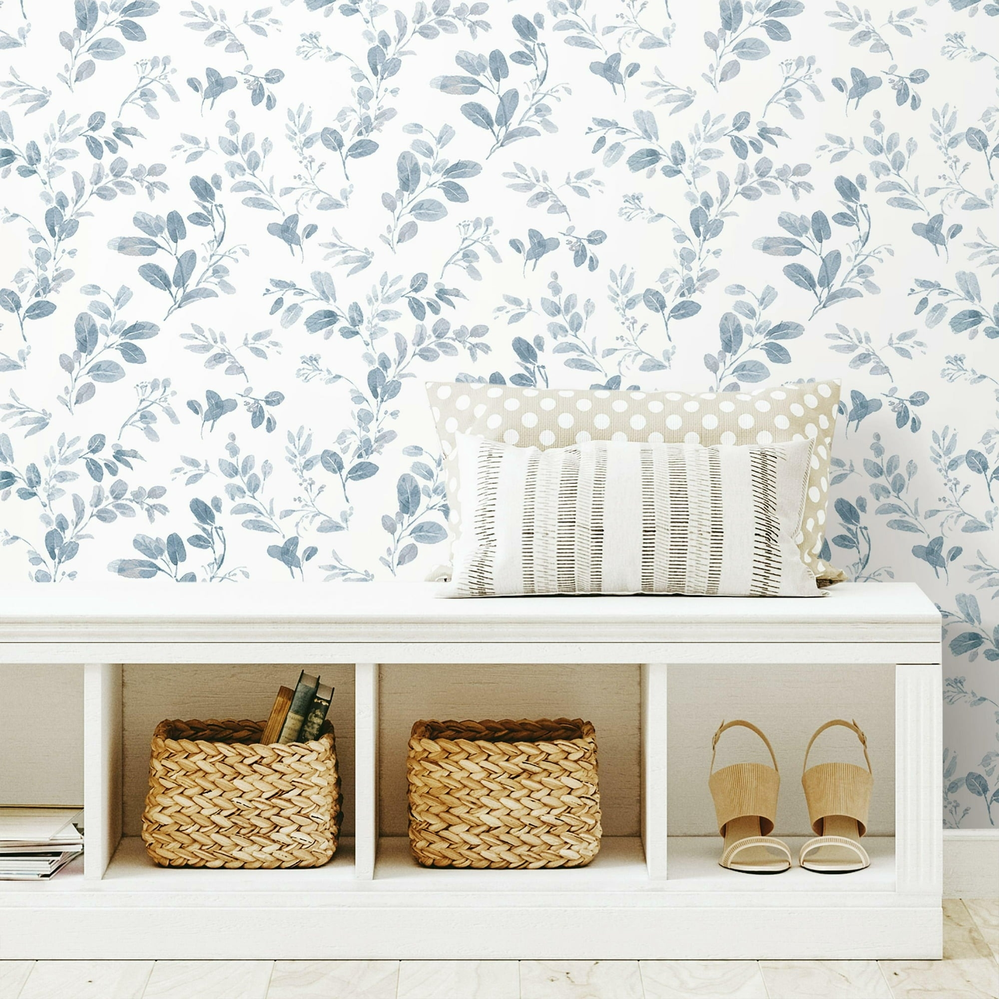 A white bench with storage baskets, books, and cushions against a floral wallpaper