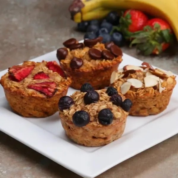 Four oatmeal muffins on a white plate, each topped with fruits and nuts, with fresh fruit in the background
