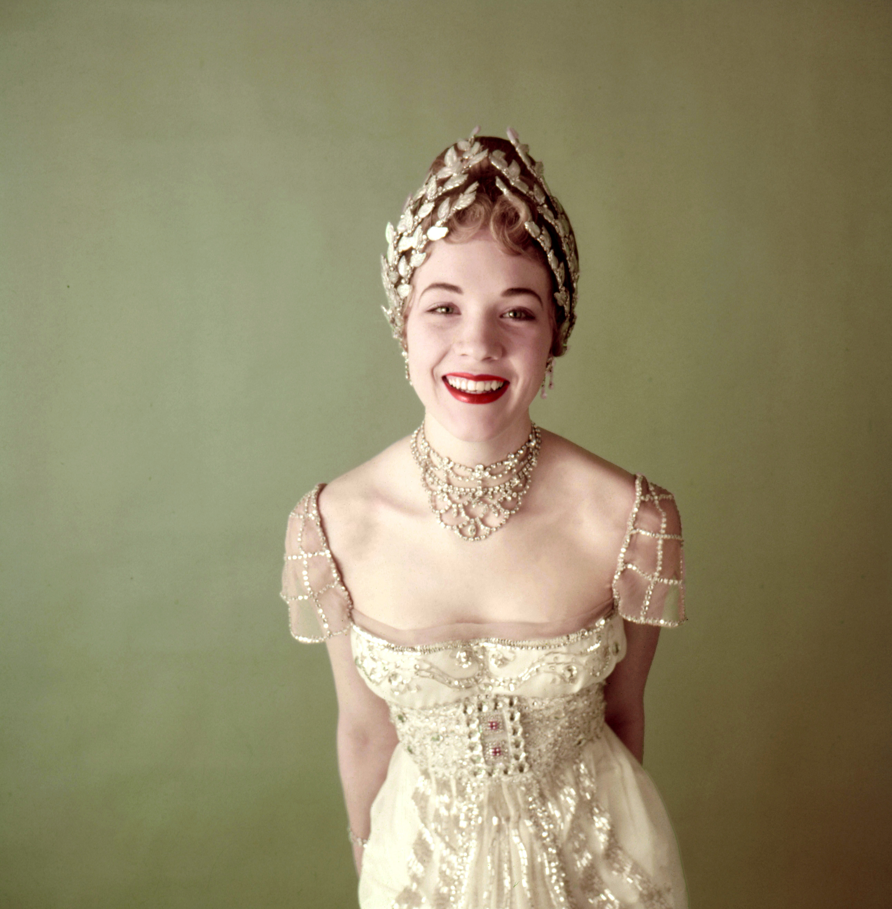 Smiling and wearing a headpiece, multistrand choker and a bejeweled, short-sleeved dress