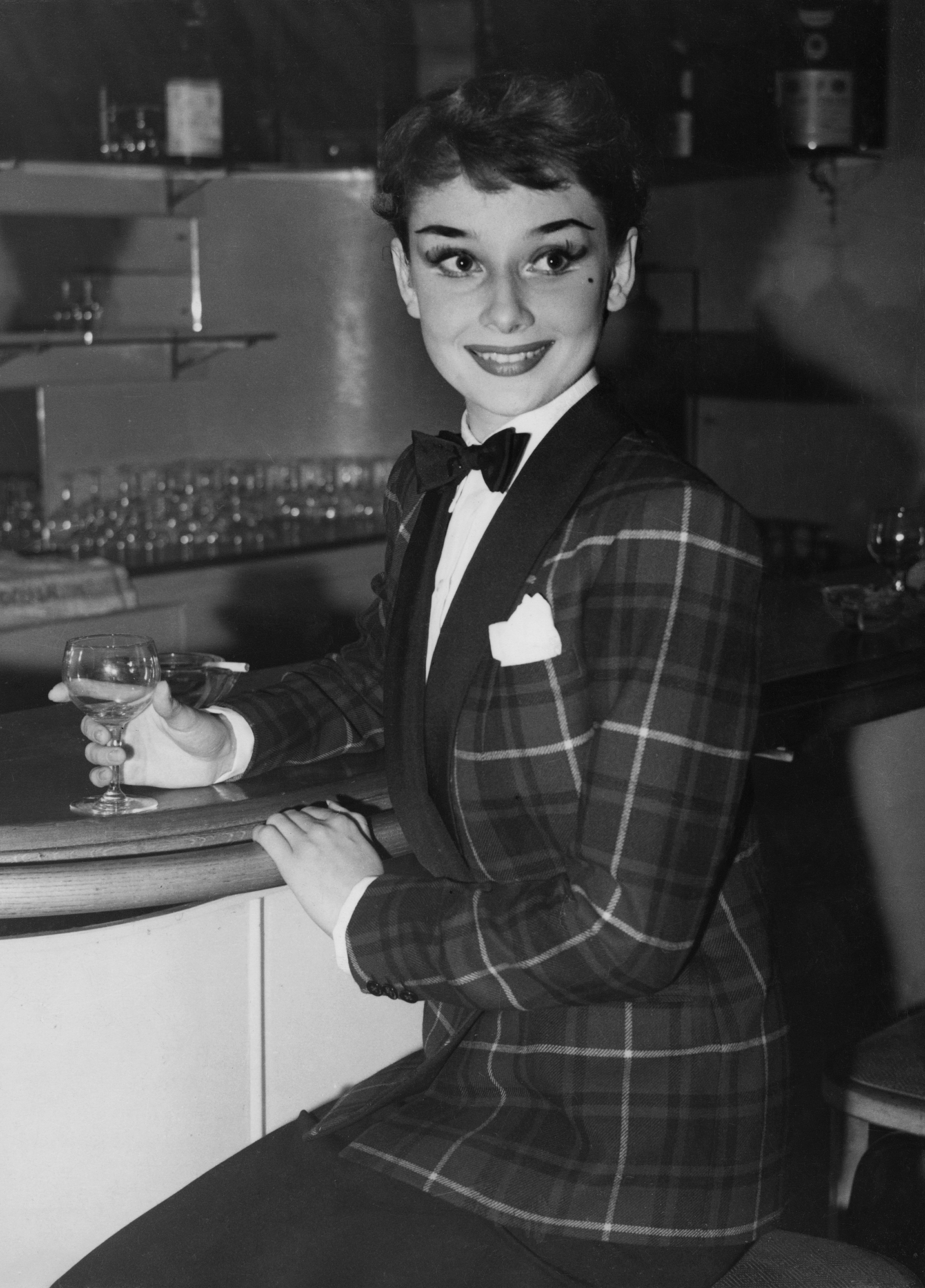 Smiling at a counter in a plaid jacket and bow tie and holding a wine glass