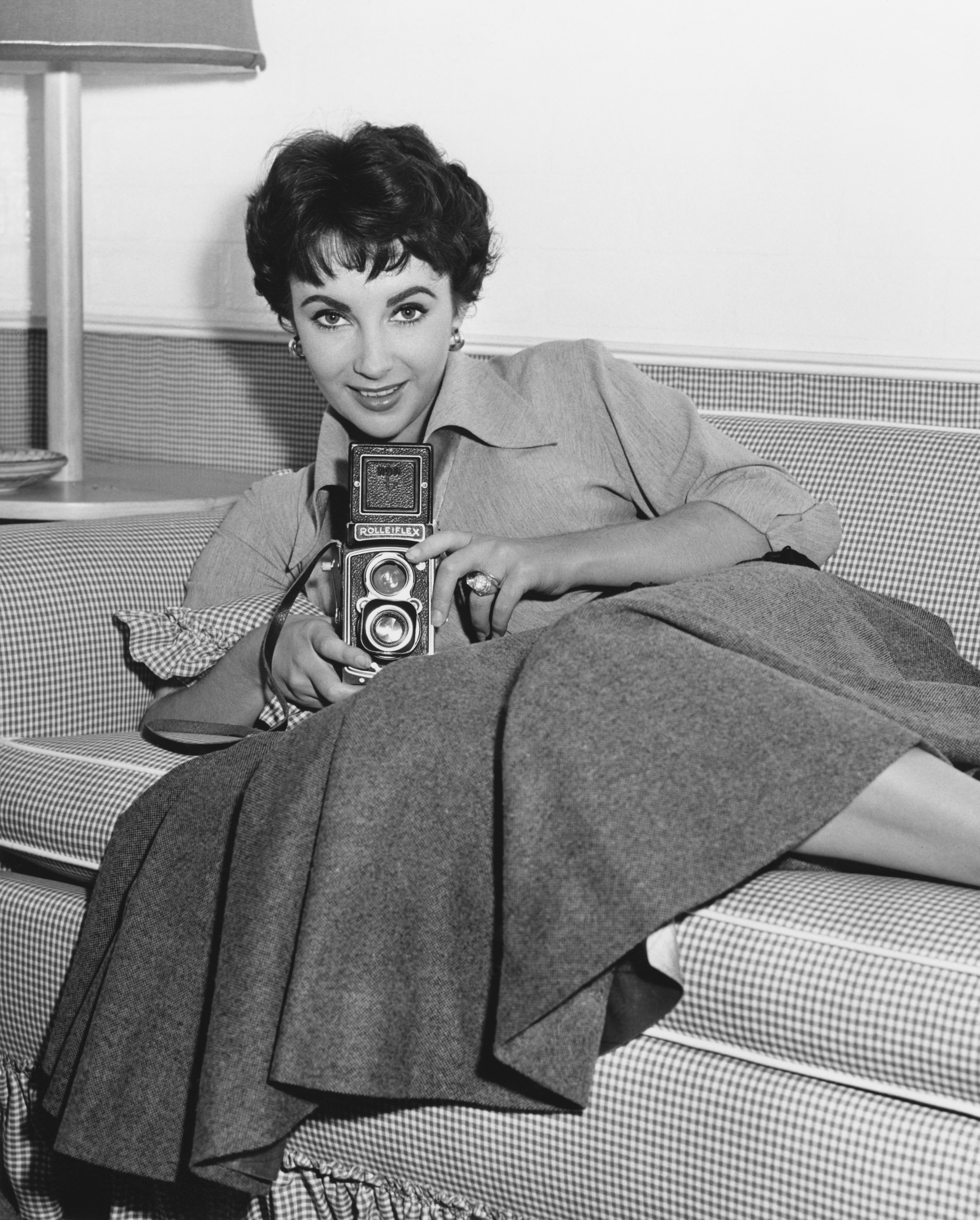 Smiling with a short haircut, wearing a flared dress, and holding a camera while sitting on a couch
