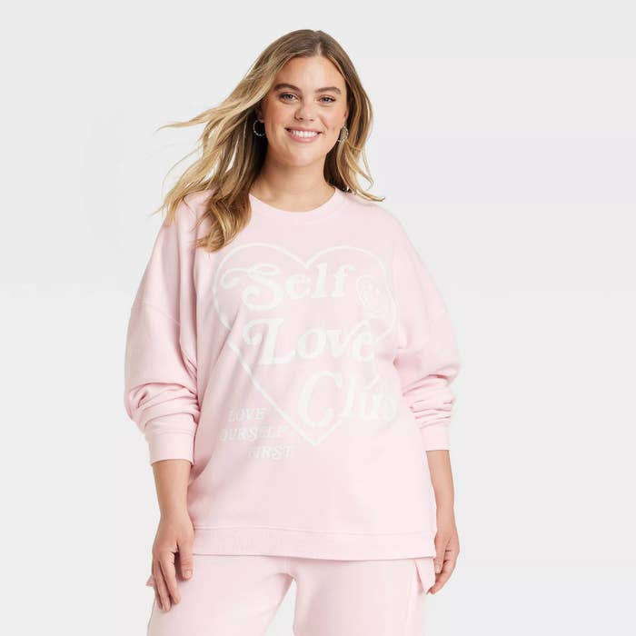 A model wearing the light pink sweatshirt with a graphic print that reads Self Love Club, featuring a crewneck and long sleeves