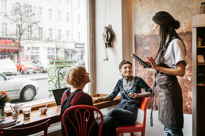 Two young women in a bright café are ordering something talking to a waitress