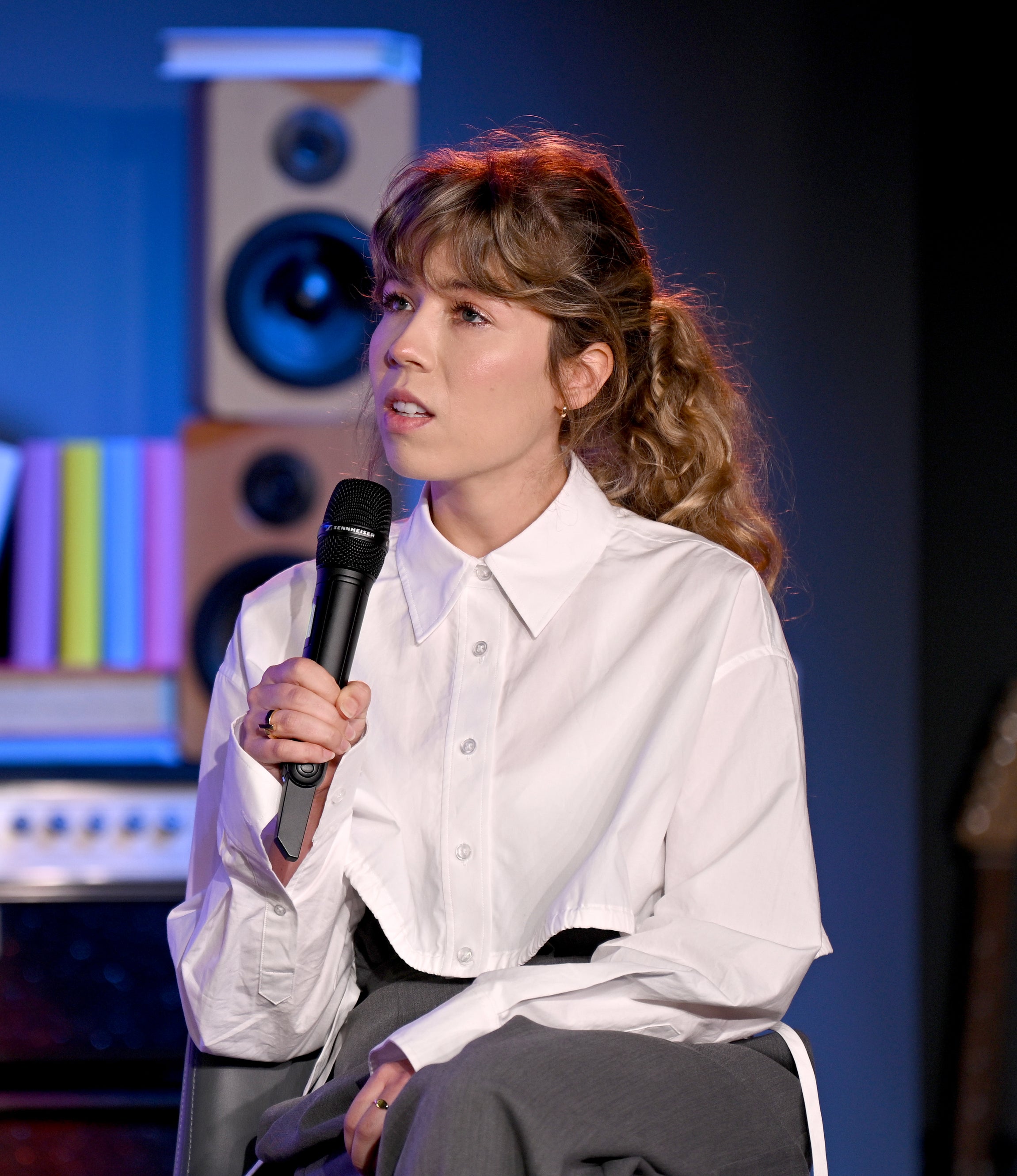 Jennette McCurdy sitting and speaking into a microphone, dressed in a shirt and dark pants, with a background of stacked books