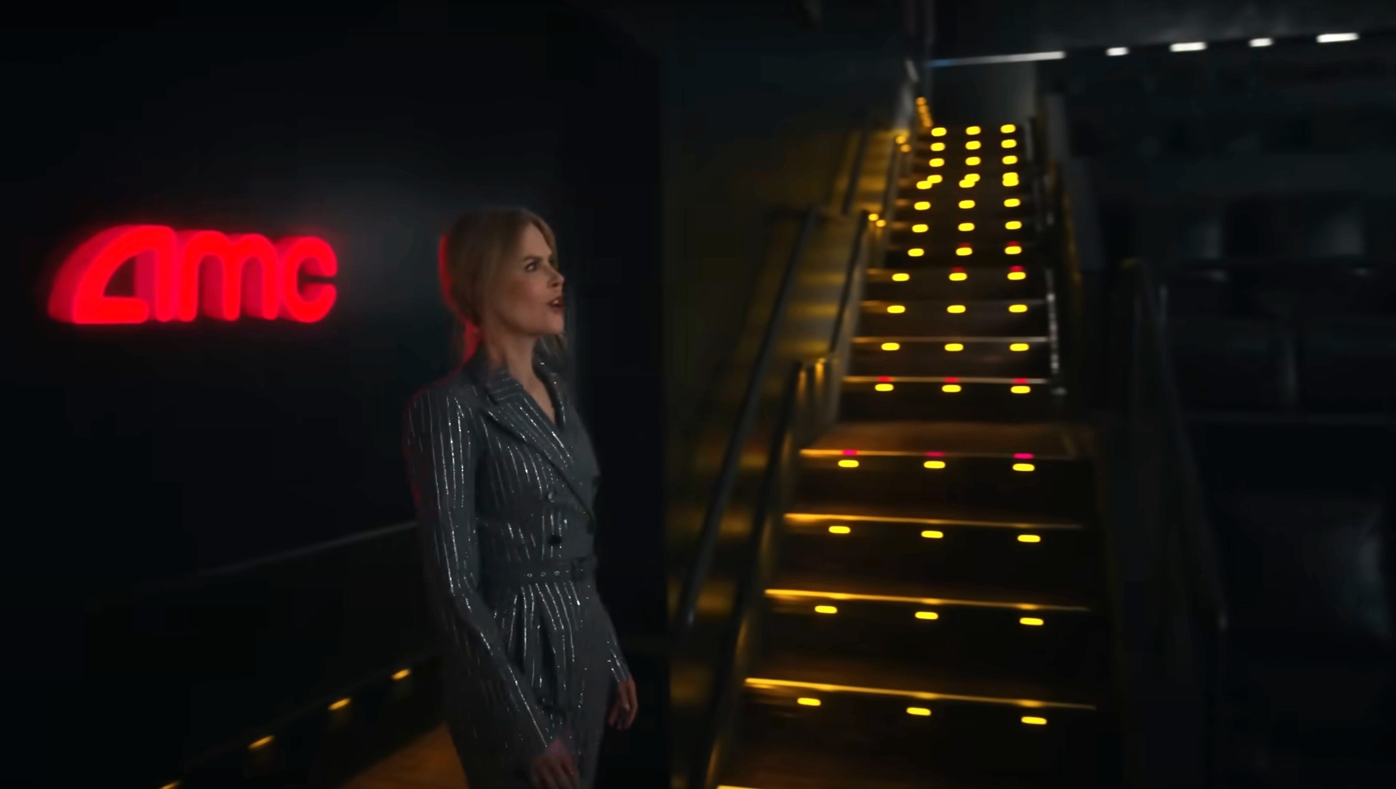 Nicole Kidman in a sparkling suit stands in a dimly lit AMC theater aisle beside illuminated stairs