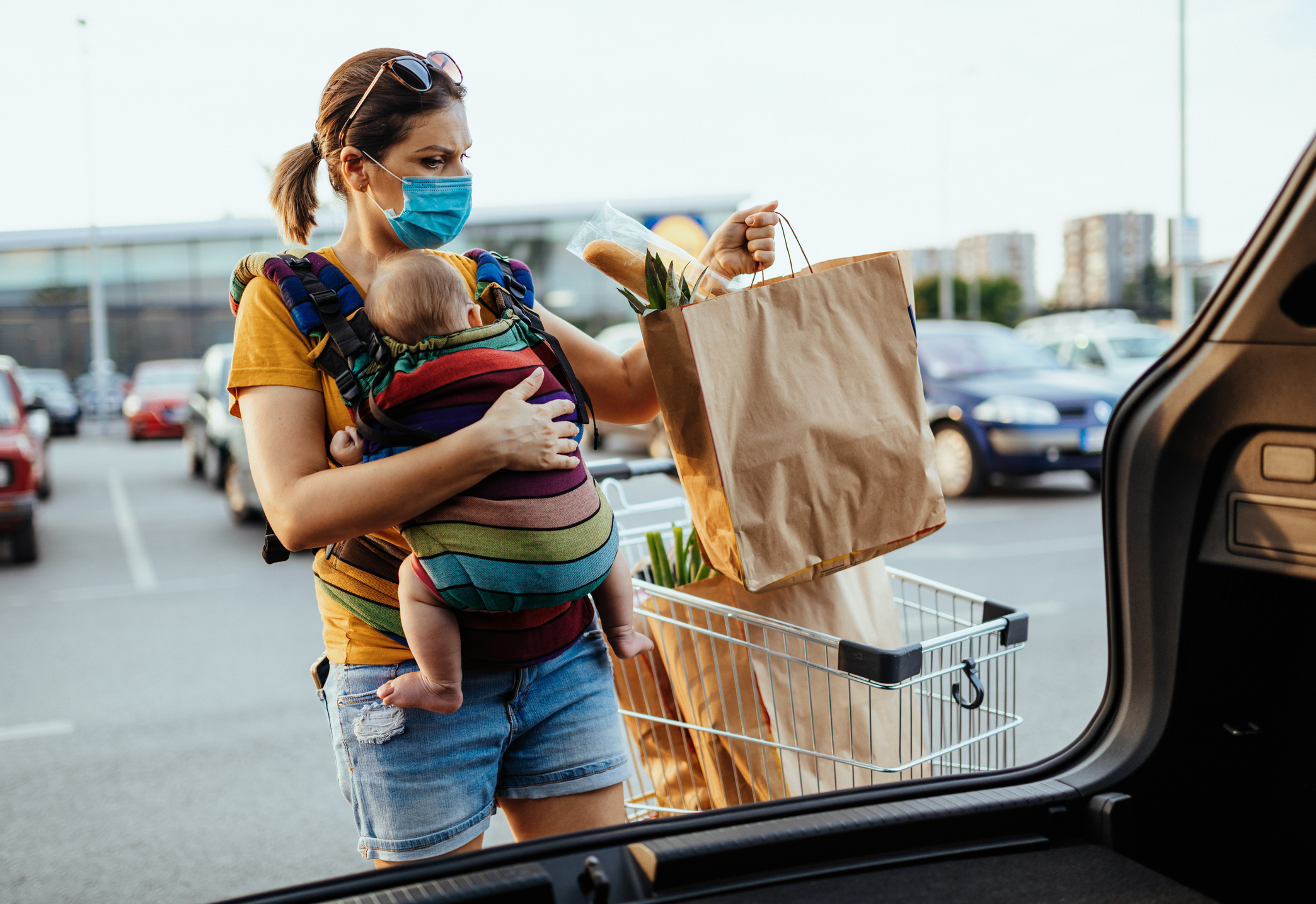 Woman wearing a mask unloading groceries, with a baby in a carrier on her chest