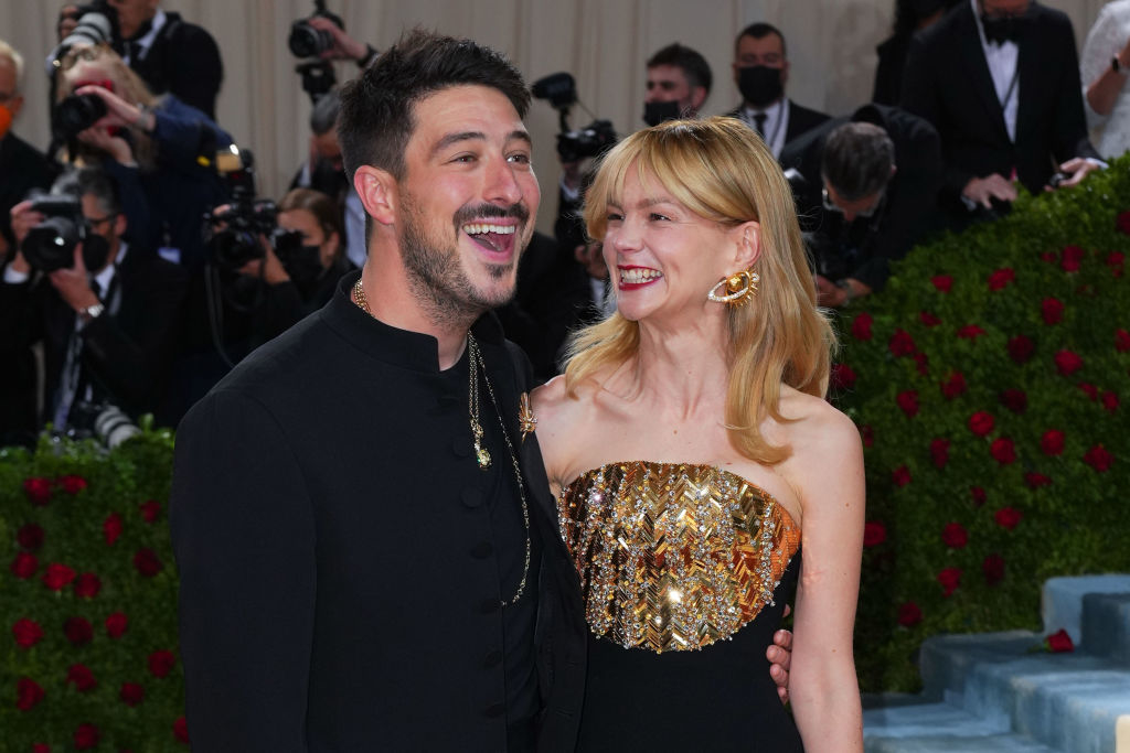 Marcus wearing a suit smiling next to Carey, who is wearing a sequined dress, on the red carpet. Photographers in the background