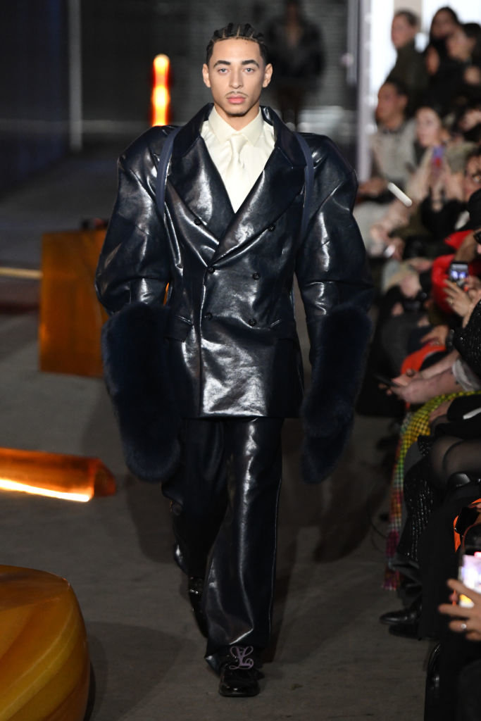 Julez walking on runway wearing oversized black jacket with fur trim and wide-leg trousers