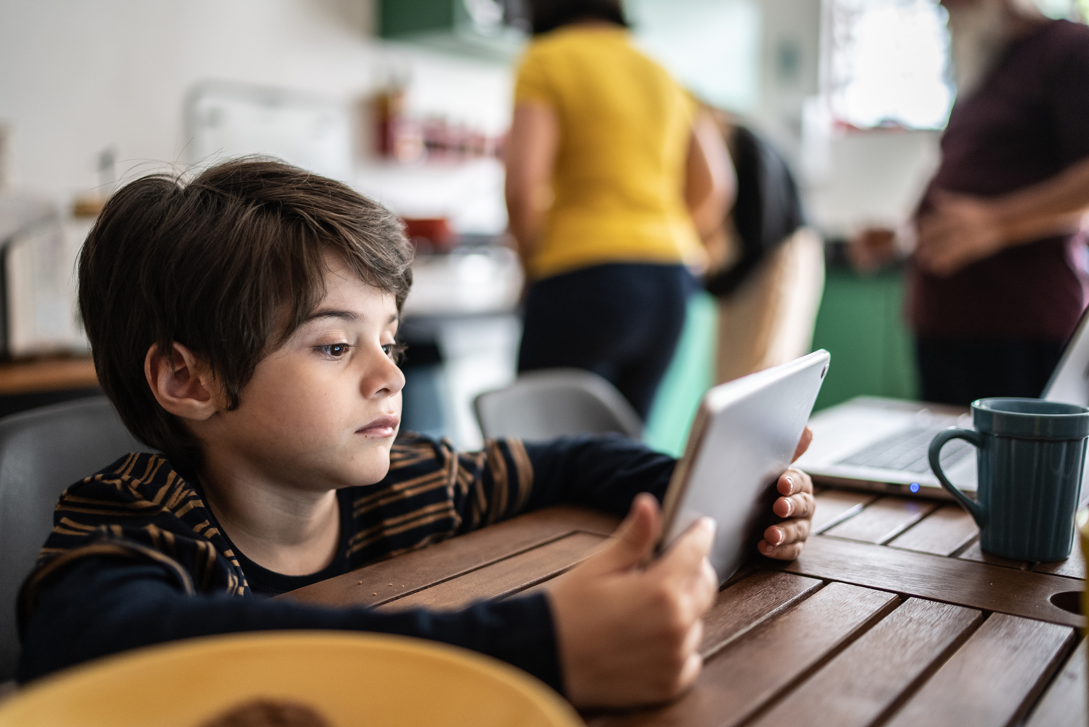 Child at a table holding a tablet with adults in the background