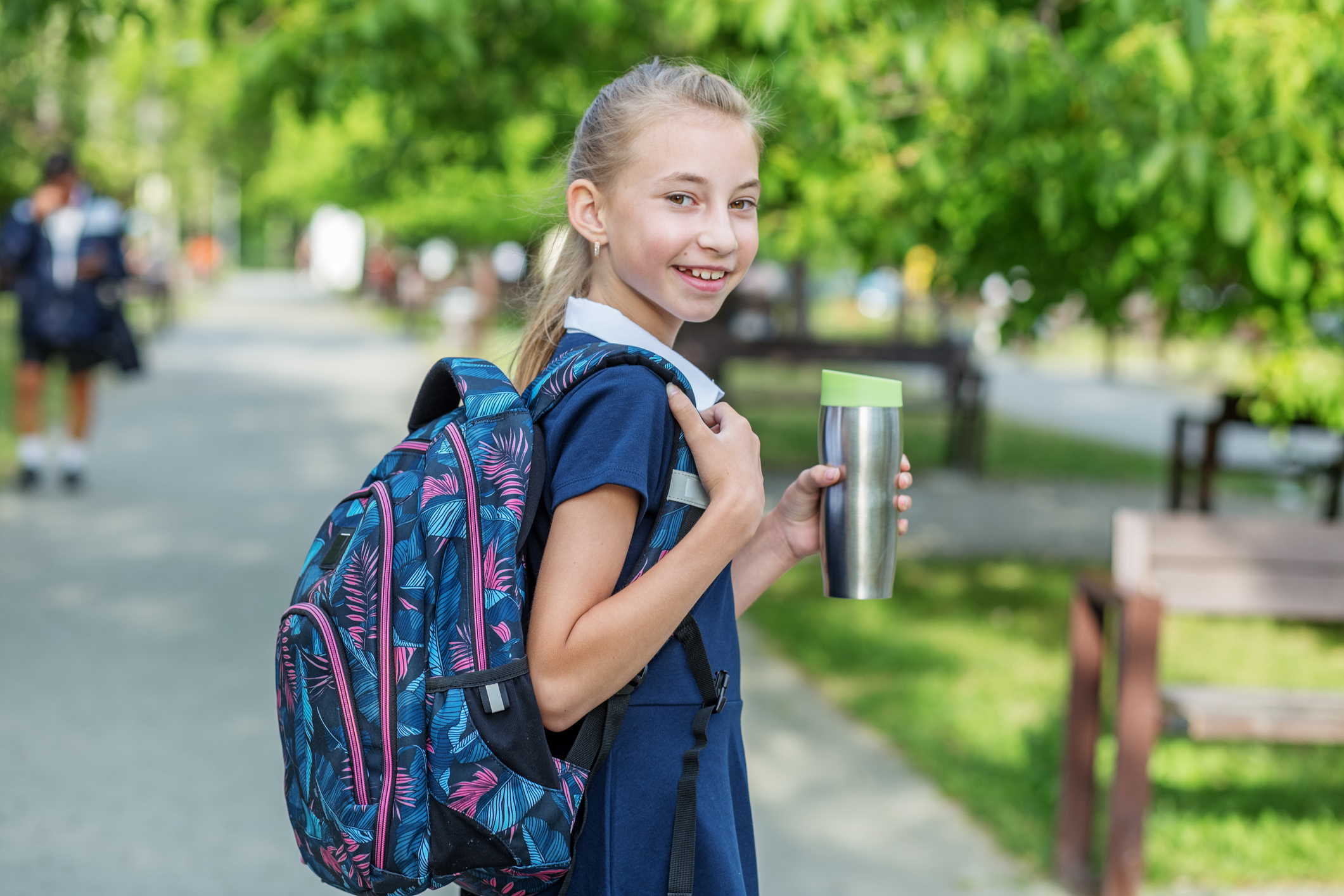 Girl with backpack smiling at camera, holding a thermos, standing on a school path