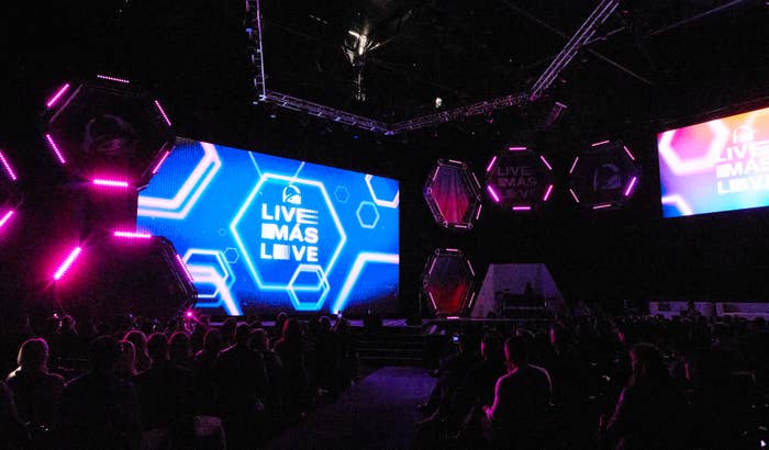 Audience looking at stage with dynamic lighting and screens displaying &quot;Live Más Live&quot; at an event