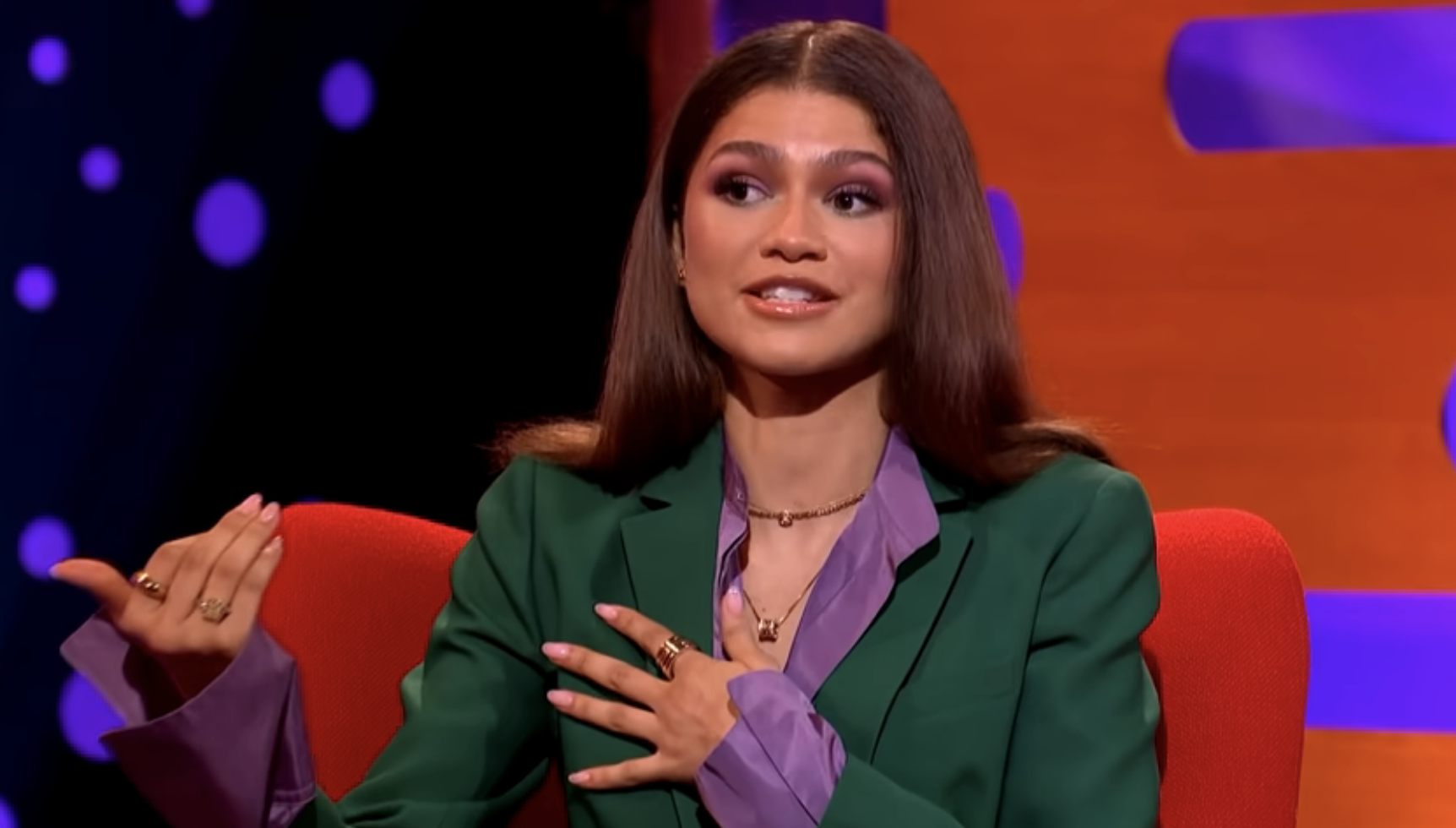 Zendaya gestures while speaking, wearing a green blazer and purple blouse, seated on a talk show set