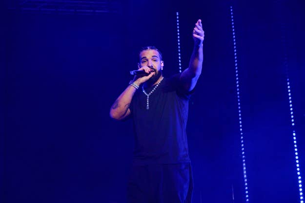 Drake performs onstage, wearing a black T-shirt and pants, with one hand raised and a microphone in the other