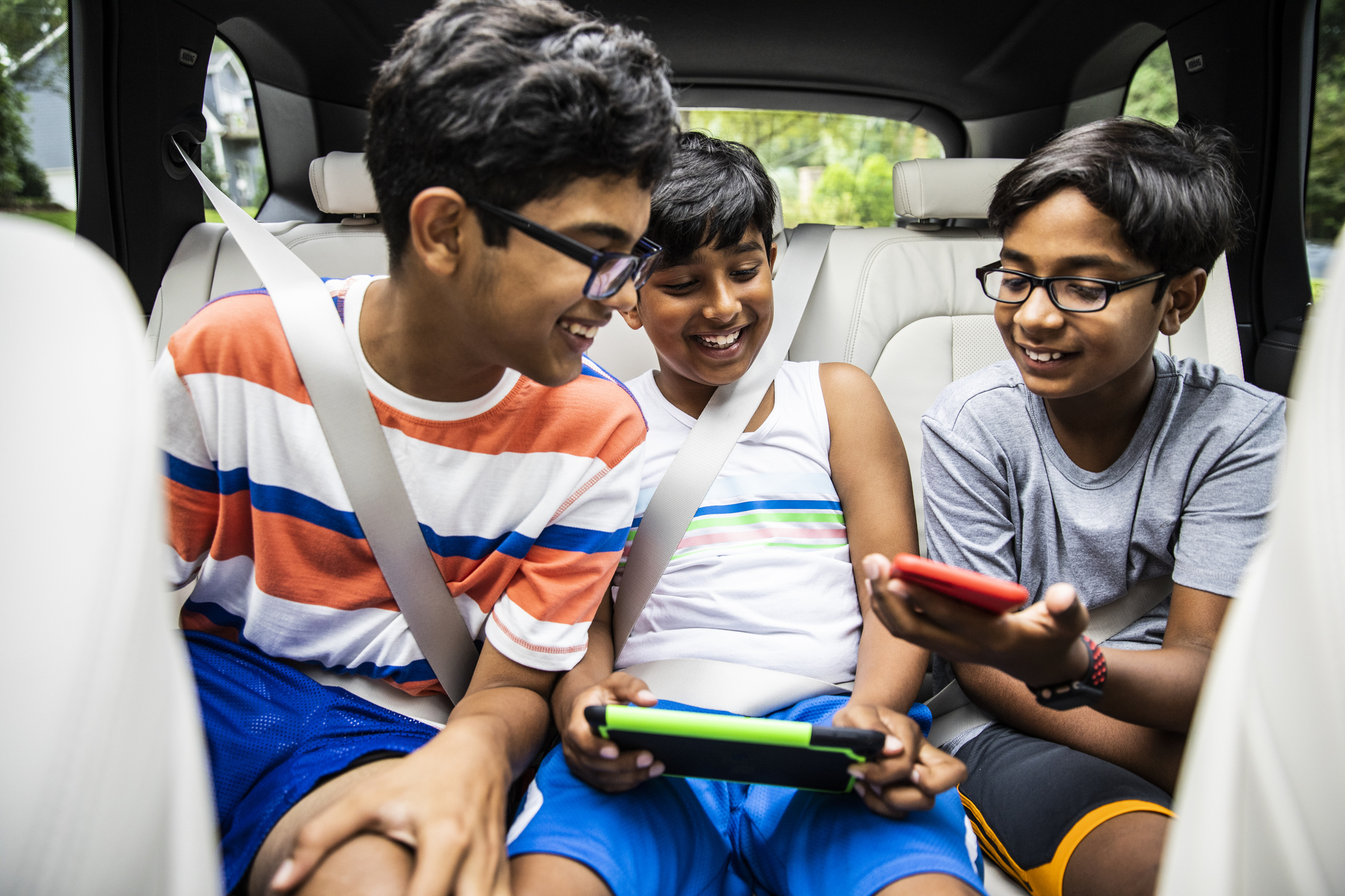Three siblings in a car use hand-held gaming devices, laughing and smiling
