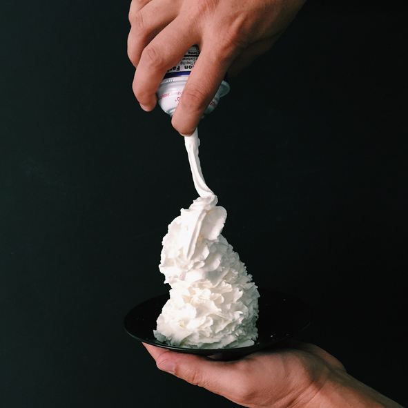Whipped cream being dispensed onto a dessert in a bowl held by a hand against a dark background