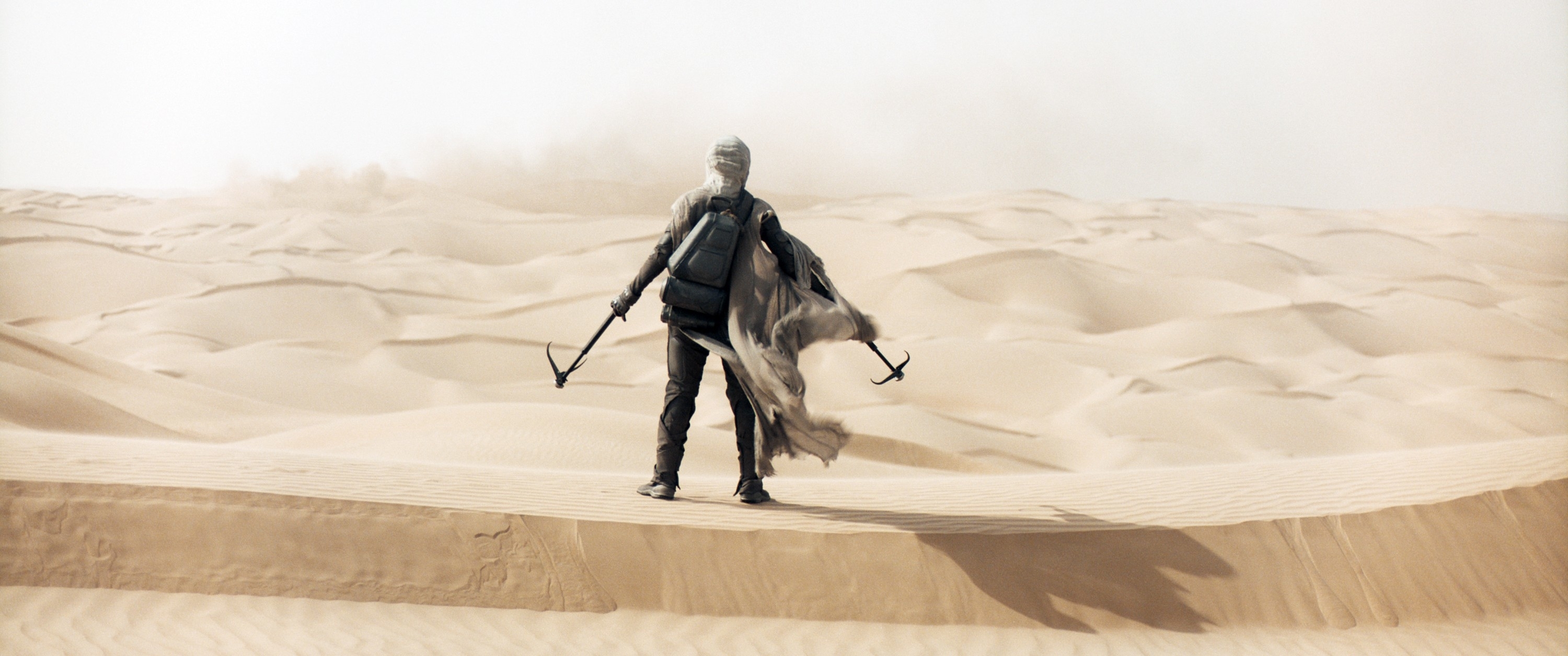Character in futuristic armor stands atop a sand dune, holding weapons, in a desert landscape