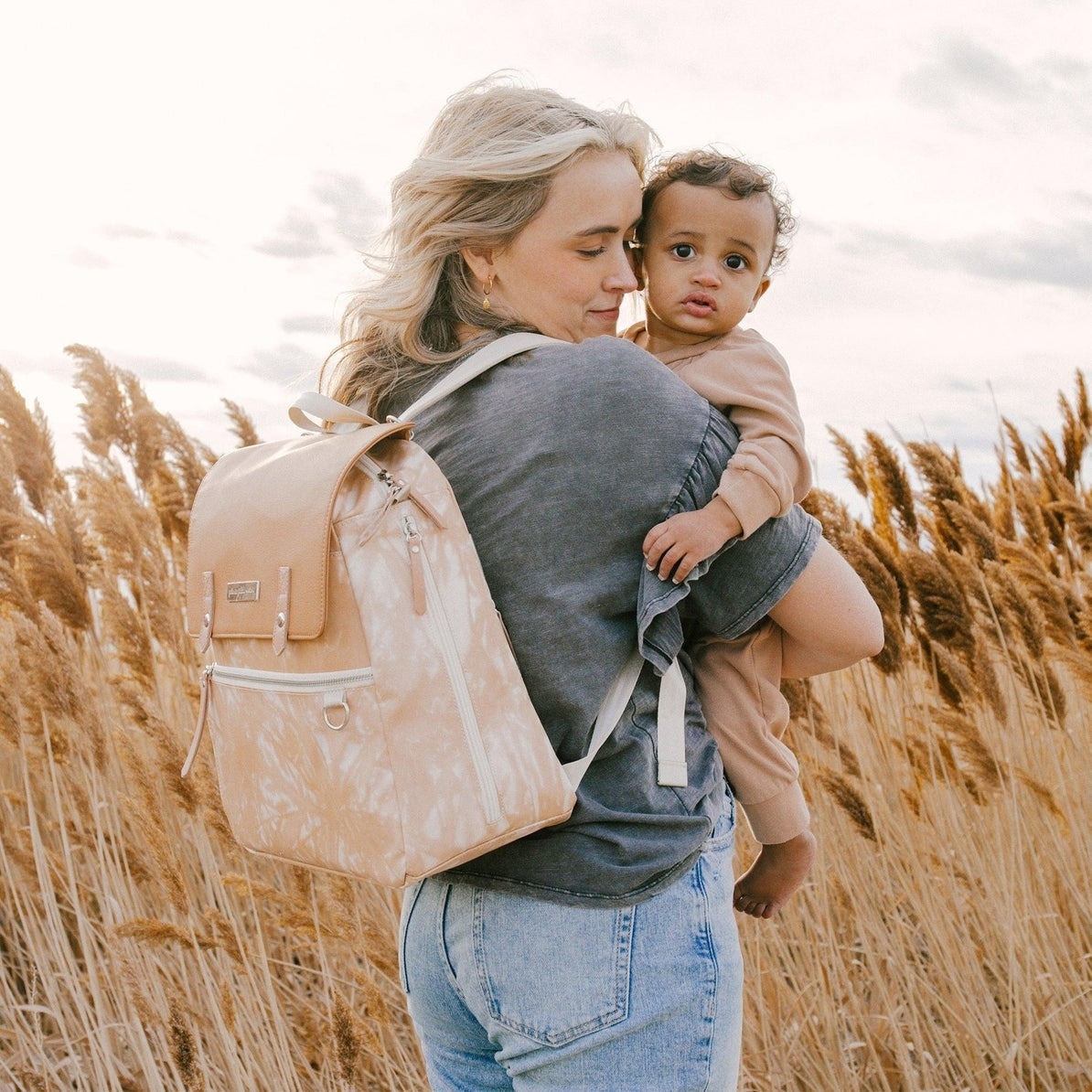 Model holding a toddler in a field, both looking toward the camera, with the taupe backpack on her shoulder