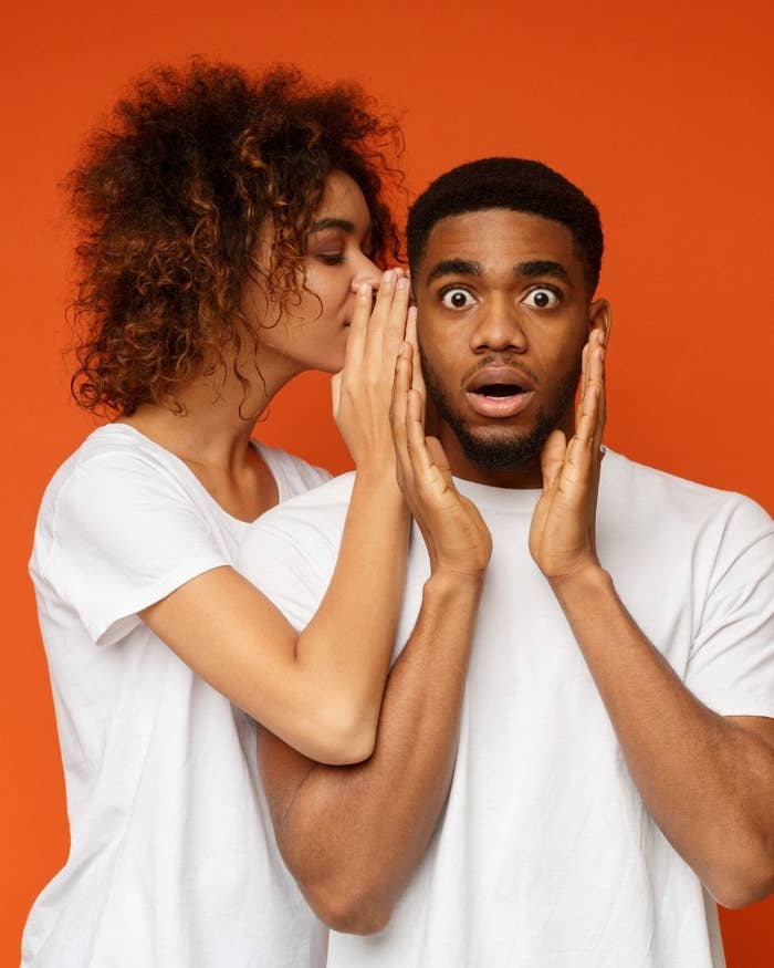 Woman whispering to surprised man, both wearing white tees against an orange backdrop