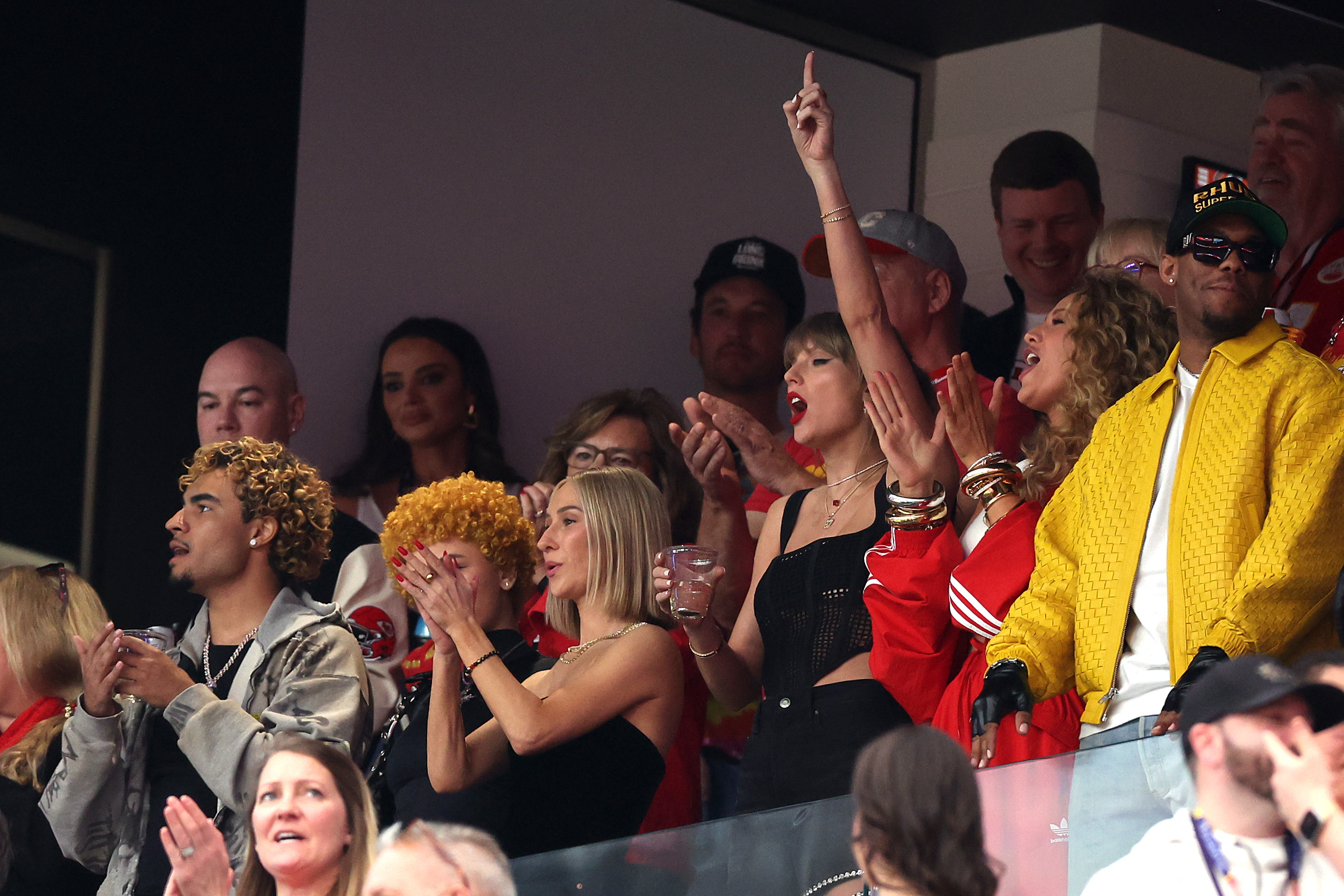 Group of people in a stadium box cheering, with Taylor pointing upward, expressions of excitement
