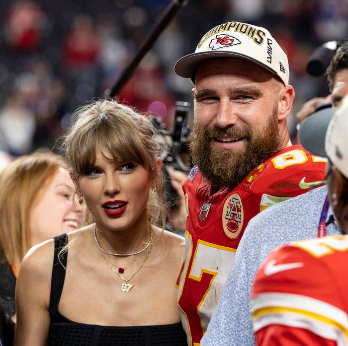 Taylor Swift stands with Travis Kelce and a player with text "Hardman 17" on a sports field. Kelce wears a cap and sports jersey