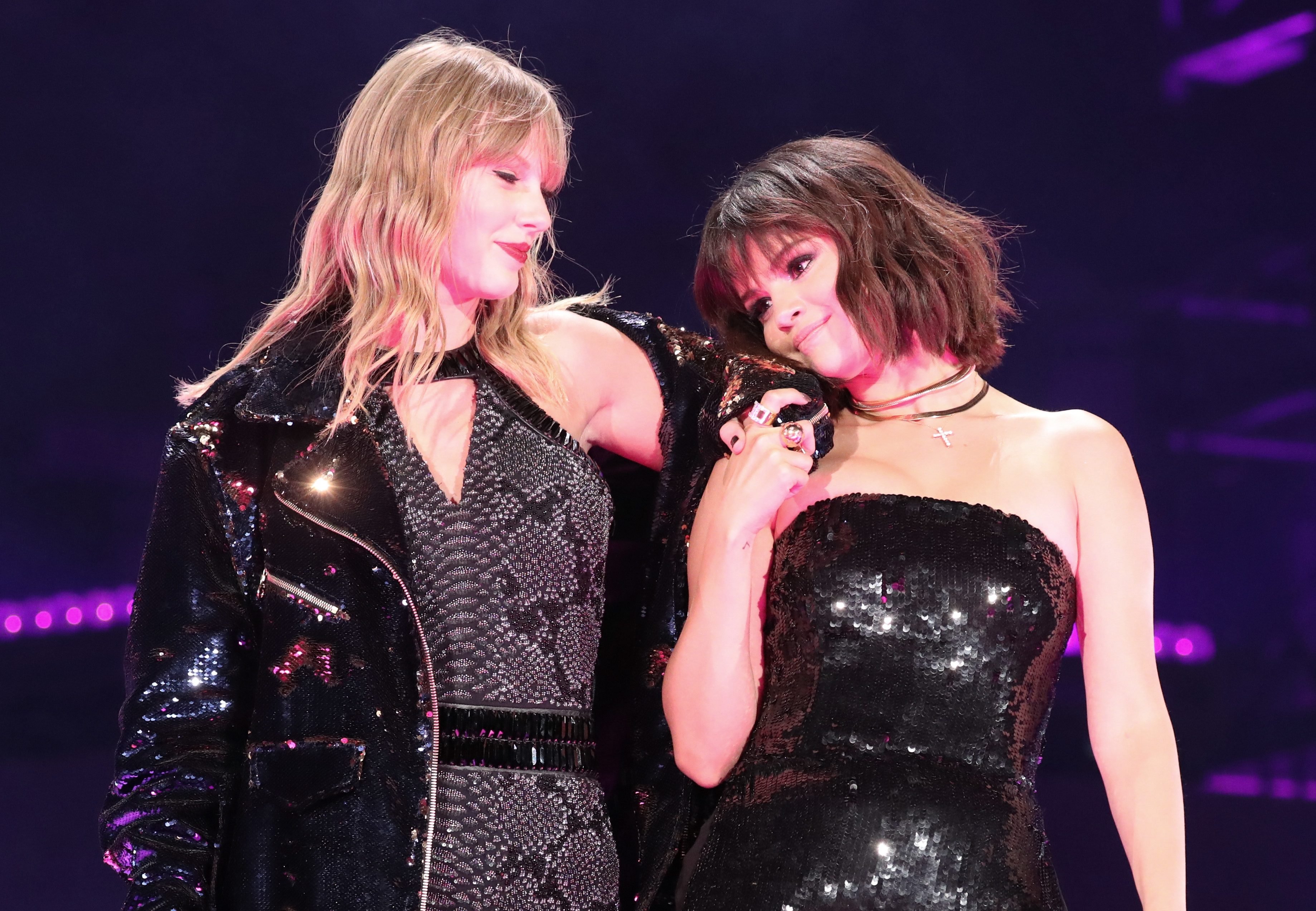 Taylor Swift and Selena Gomez on stage, wearing sequined outfits and smiling at each other