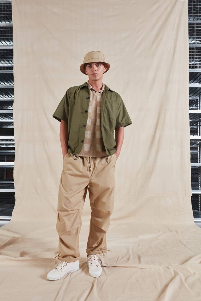 Person in a bucket hat, shirt over sweater, khaki pants, and white sneakers, posing with hands in pockets