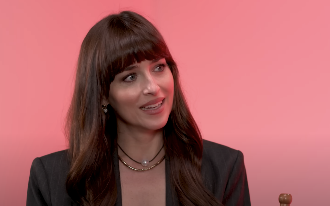 Dakota, with bangs and layered necklace, speaks in front of a pink background