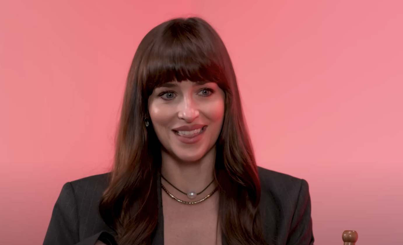 Dakota smiling in front of a pink background; she wears a chain necklace