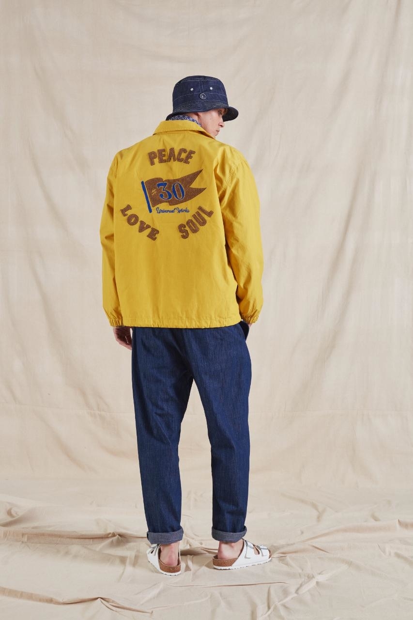 Person in a yellow jacket with &quot;PEACE LOVE SOUL&quot; on the back, denim pants, and a hat, standing against a beige backdrop