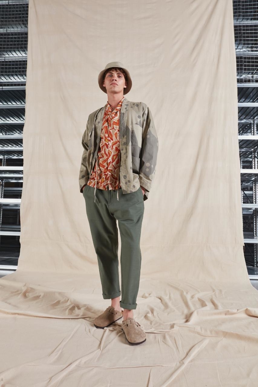 Person posing in a layered outfit with bucket hat, patterned shirt, and olive jacket