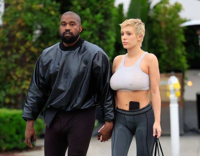 Ye in a black jacket and pants holding hands with Bianca, who&#x27;s in a gray sports crop top and leggings, both walking