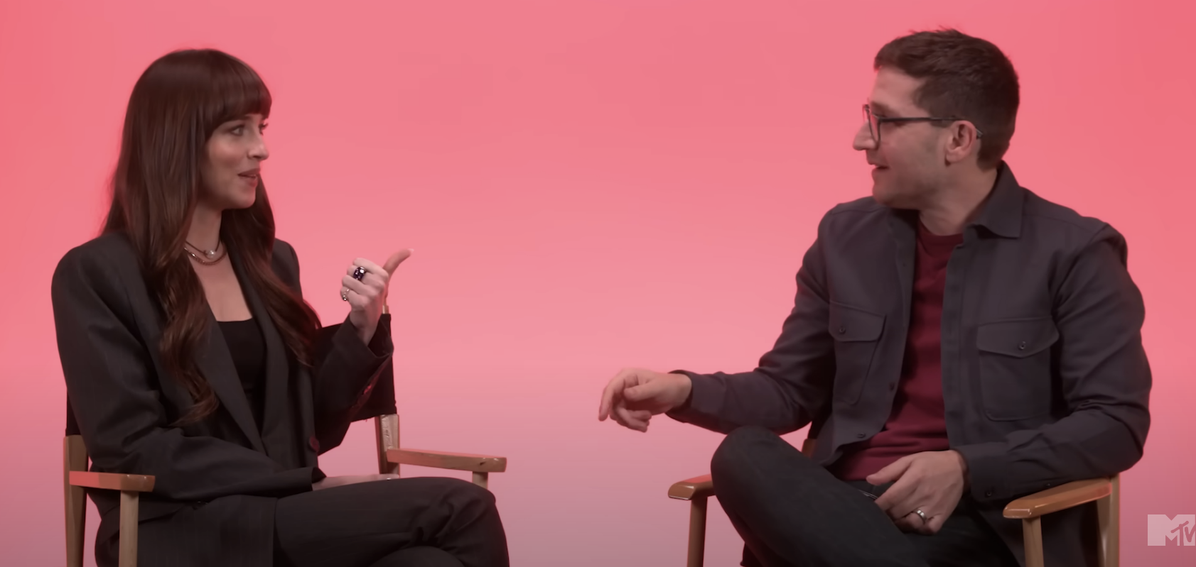 Dakota and Josh sitting, chatting, one gesturing with hand; they&#x27;re in front of a pink background