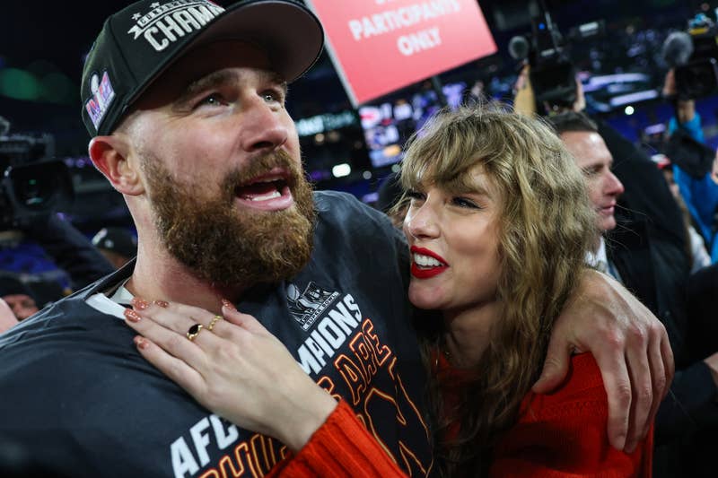 Two people embracing joyfully surrounded by cameras, man in a casual printed tee, woman in a red knit