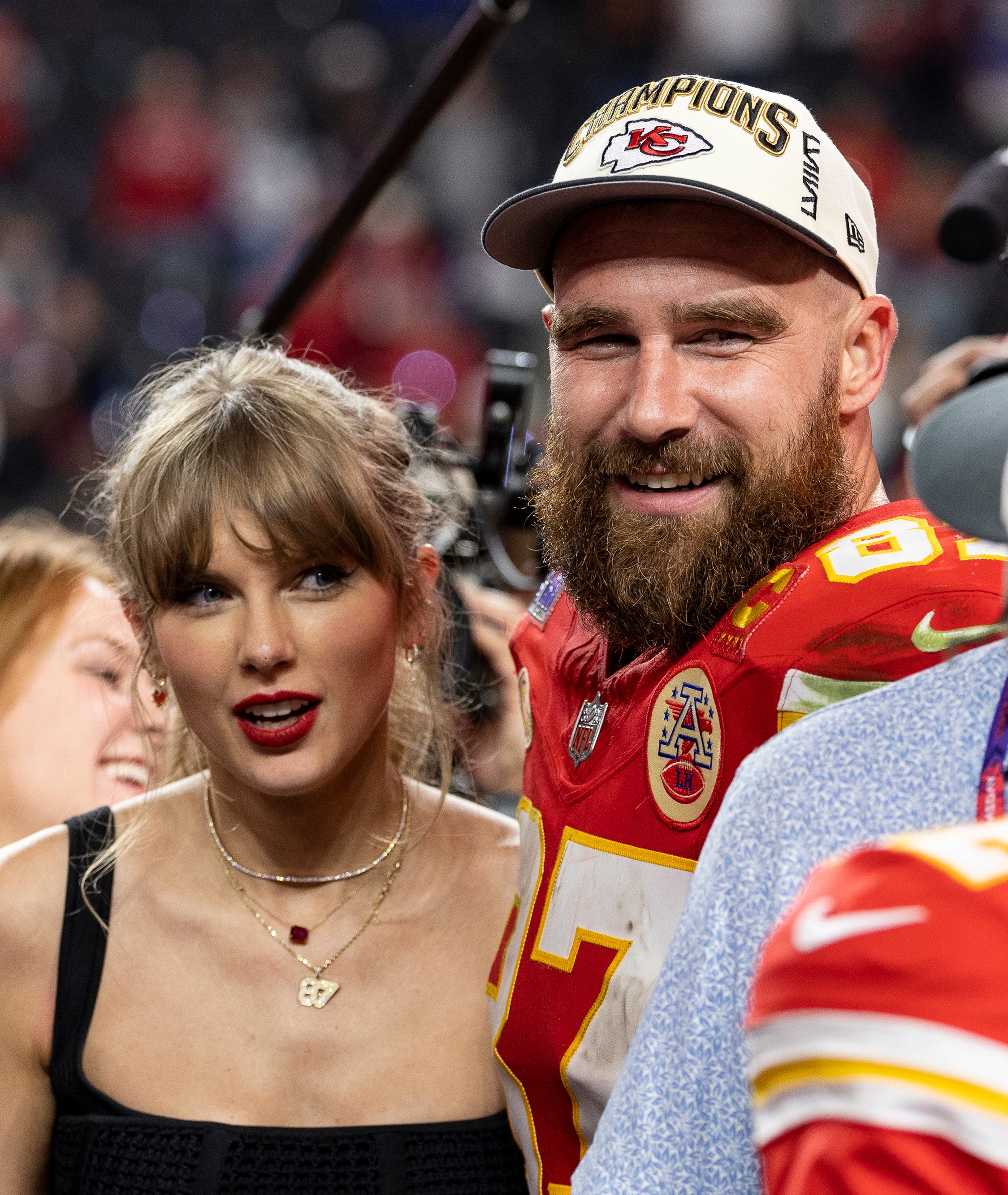 Travis Kelce and Taylor Swift smiling at an event, Kelce in Chiefs gear, Swift in dark top